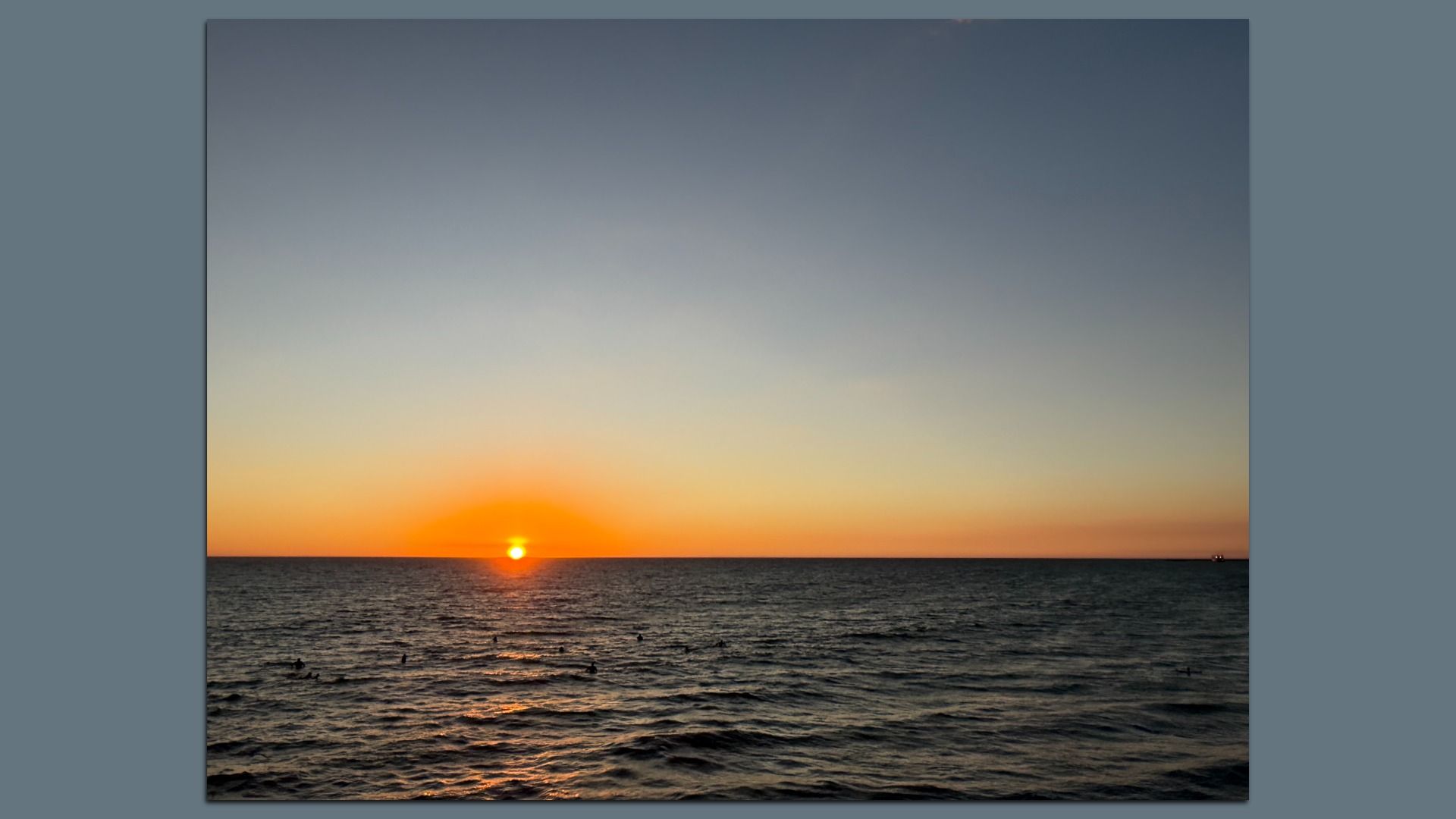 Sunset over a calm sea with an orange sun on the horizon, casting warm light on the dark blue water. The sky fades from orange-yellow near the horizon to pale blue overhead, with distant silhouettes.