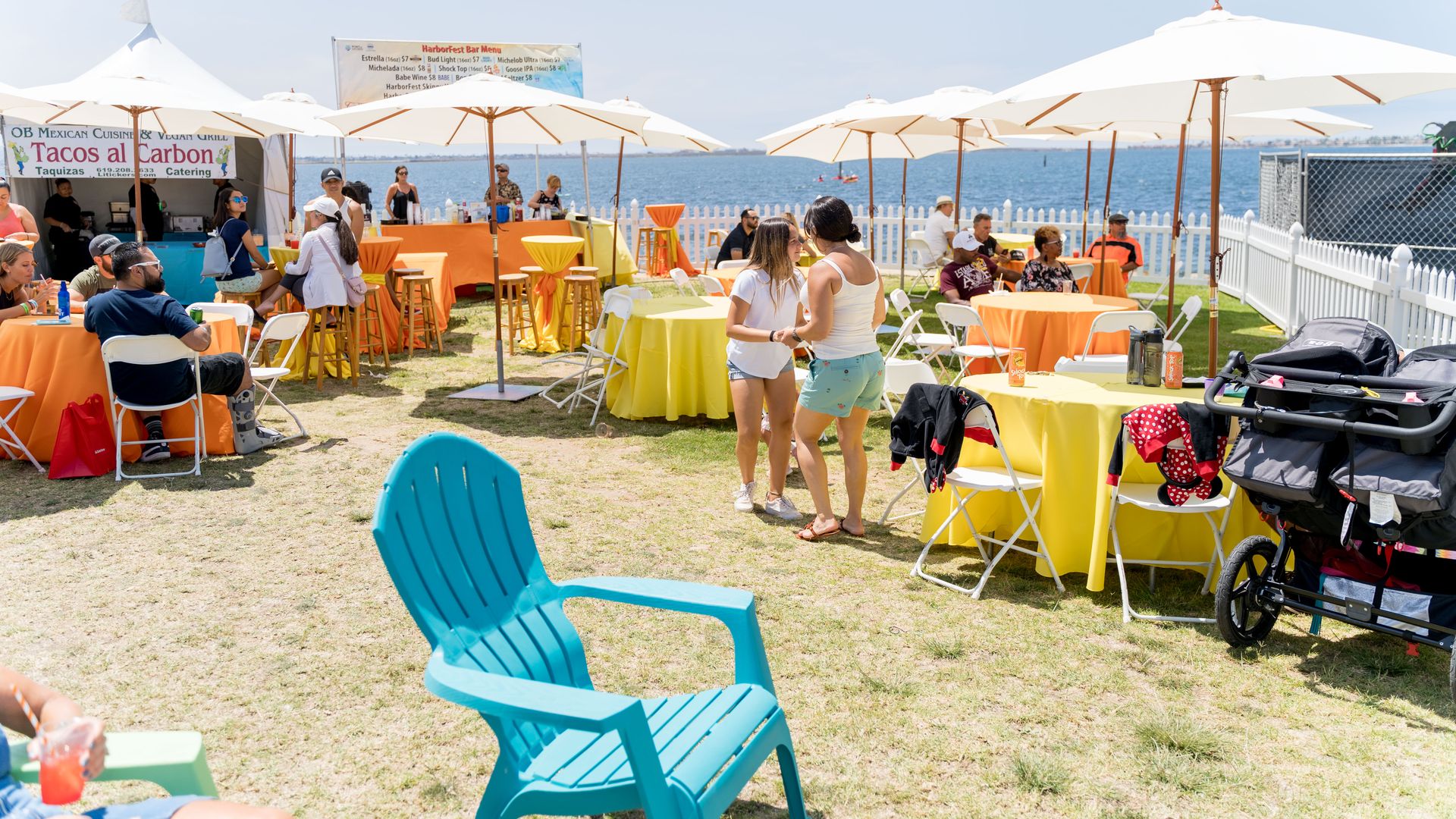 Beachfront festival with white umbrellas, orange and yellow tablecloths, and people seated at tables; a taco stand reads "Tacos al Carbon" as the blue ocean and clear sky stretch behind.