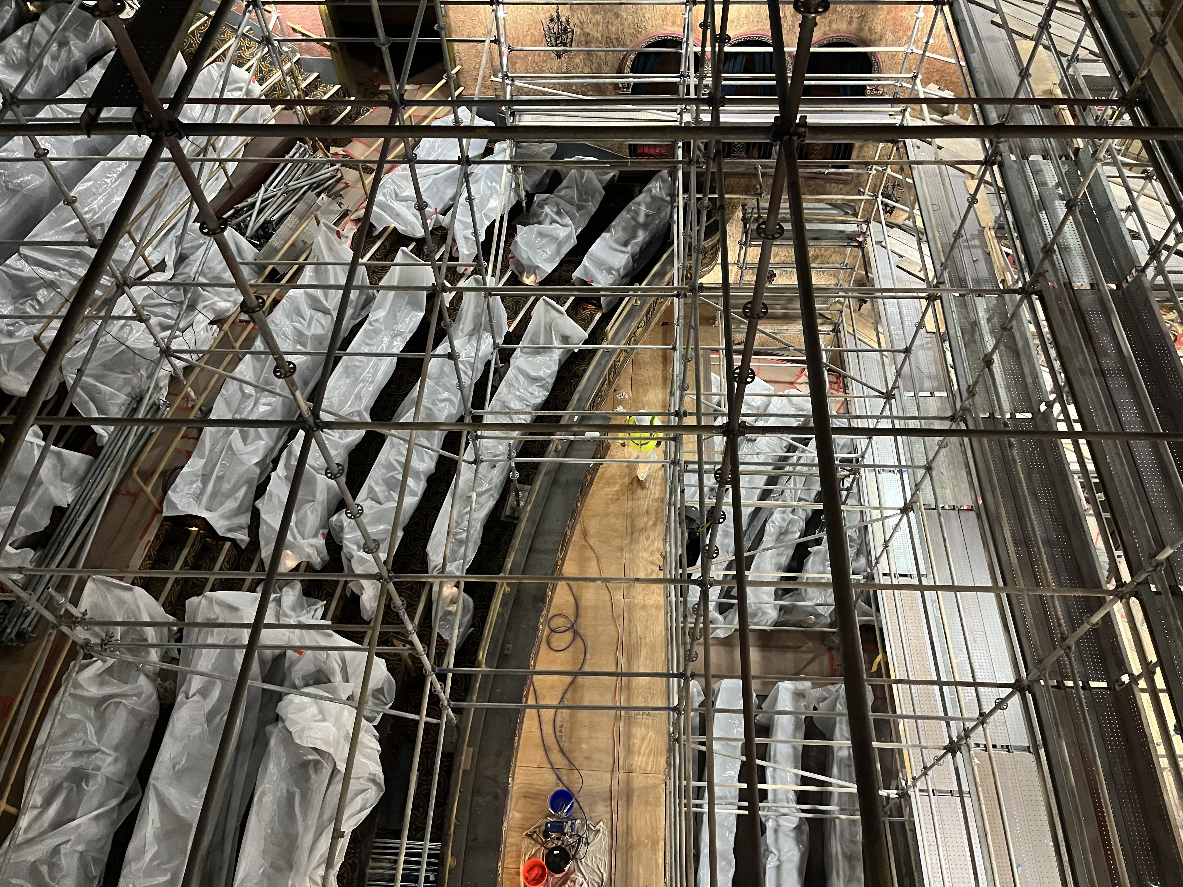 View inside a building under renovation: metal scaffolding fills the frame, plastic sheeting covers walls, and a wooden floor runs center; ornate architectural details peek through.