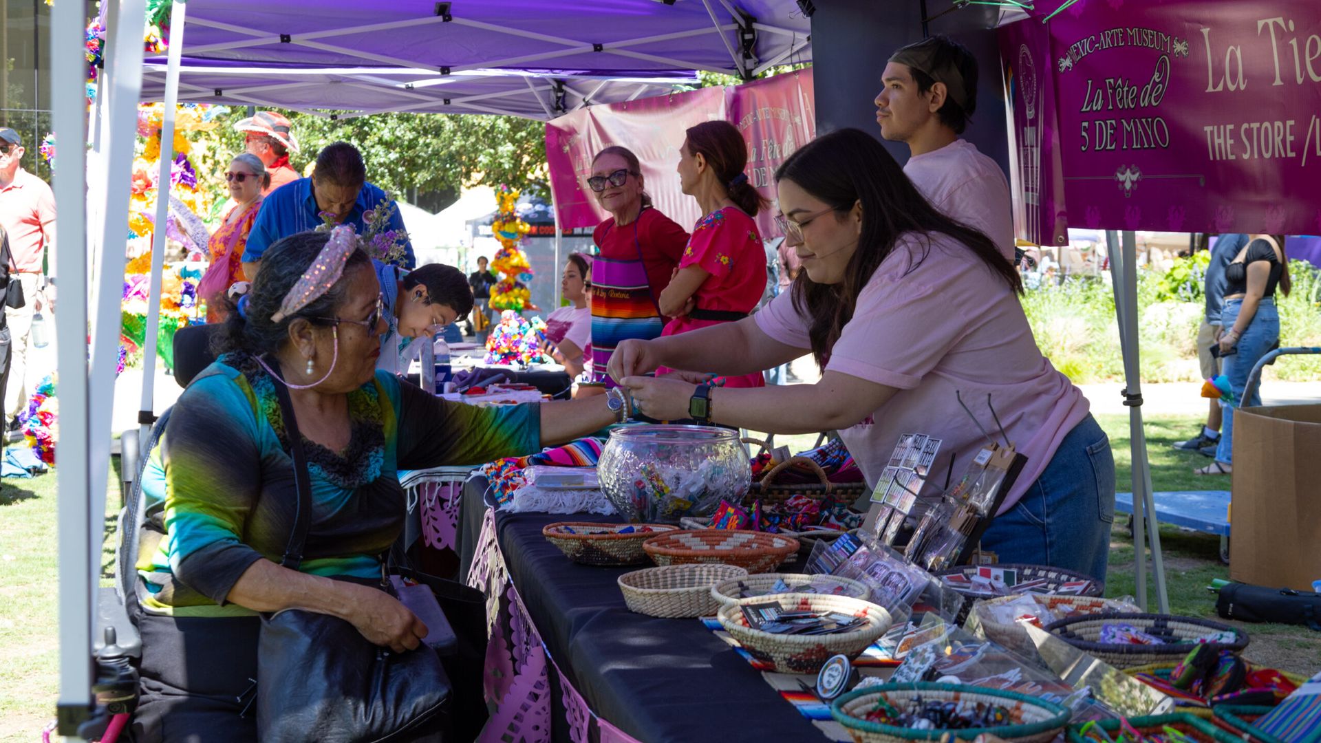 Outdoor festival market under purple tents; a vendor helps a seated woman try on colorful bracelets amid tables of woven crafts.