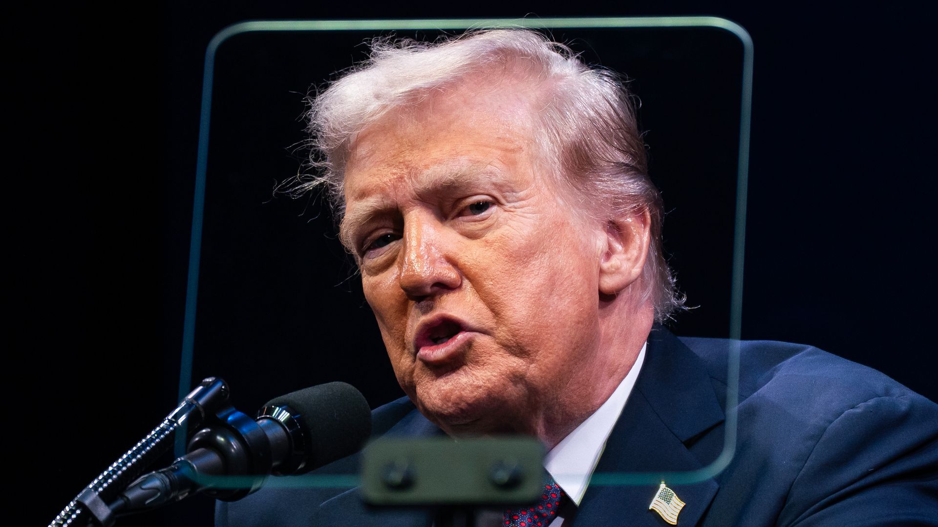 President Trump wearing a blue suit and speaking into a microphone while in front of a glass teleprompter.