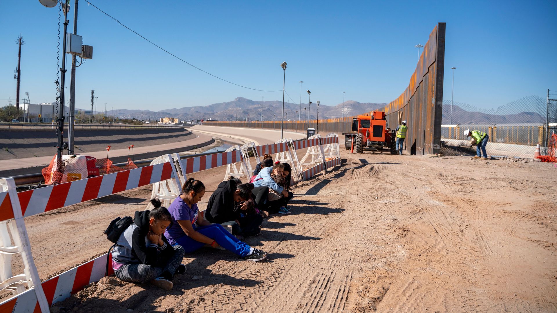 migrants wait for a transport to arrive after turning themselves into US Border Patrol by border fence under construction