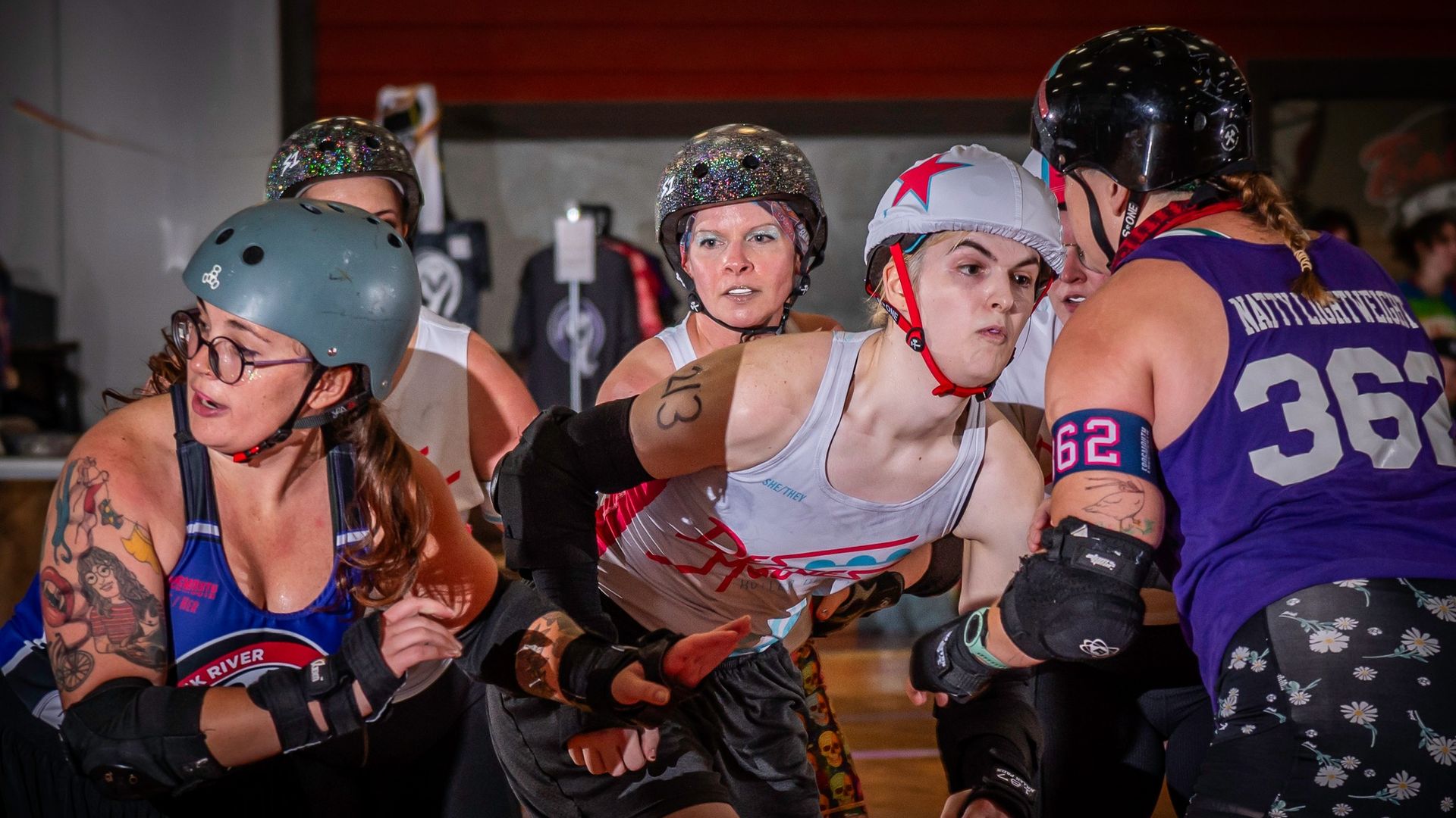 Five roller derby players in helmets and protective gear compete intensely indoors, with jerseys in white, blue, and purple, tattoos visible on arms, and focused expressions.