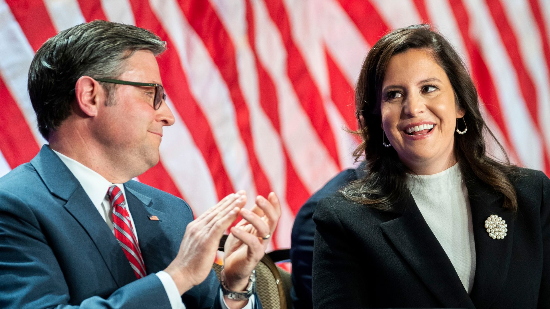 Mike Johnson wearing a gray suit and Elise Stefanik wearing a black suit sitting in front of the red and white stripes of a large American flag.