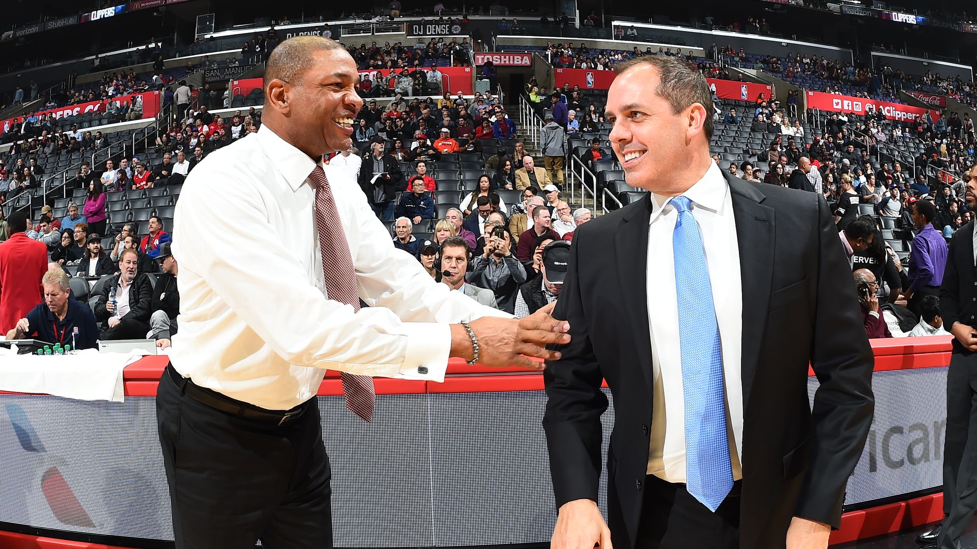 A smiling man in a shirt and tie grabs the arm another smiling man in a suit. 