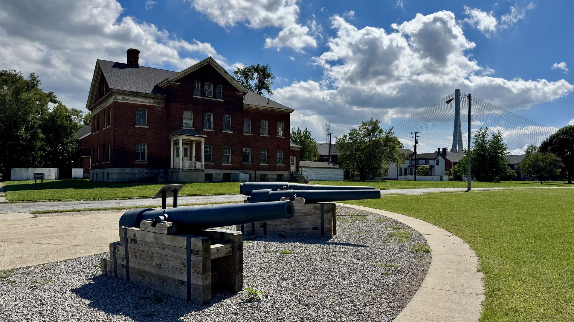 Red brick, two-story building with white trim on the left; a grassy lawn and gravel foreground with large blue pipes; a cable-stayed bridge rises on the right under a cloudy blue sky.