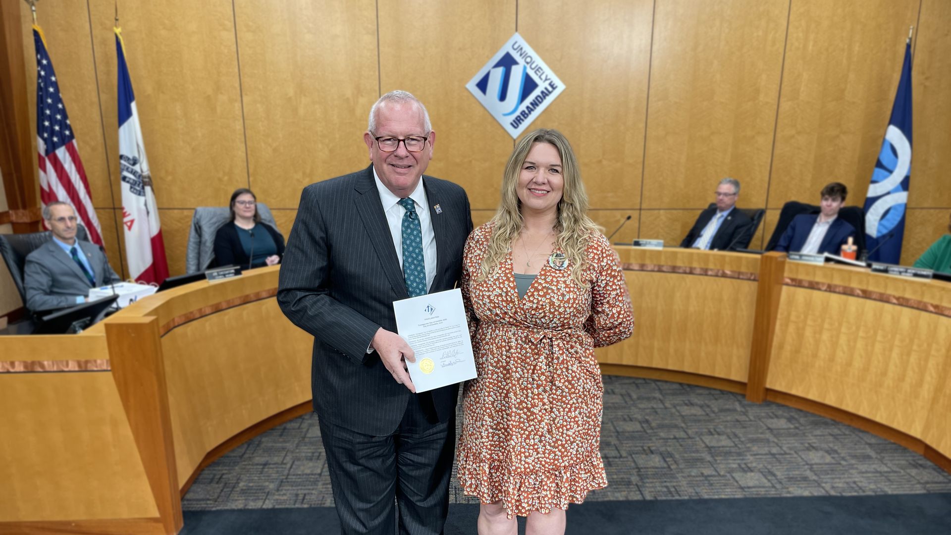 Man in a dark suit and a woman in a red floral dress stand in a wood-paneled council chamber, holding a certificate; flags and seated officials are visible in the background.