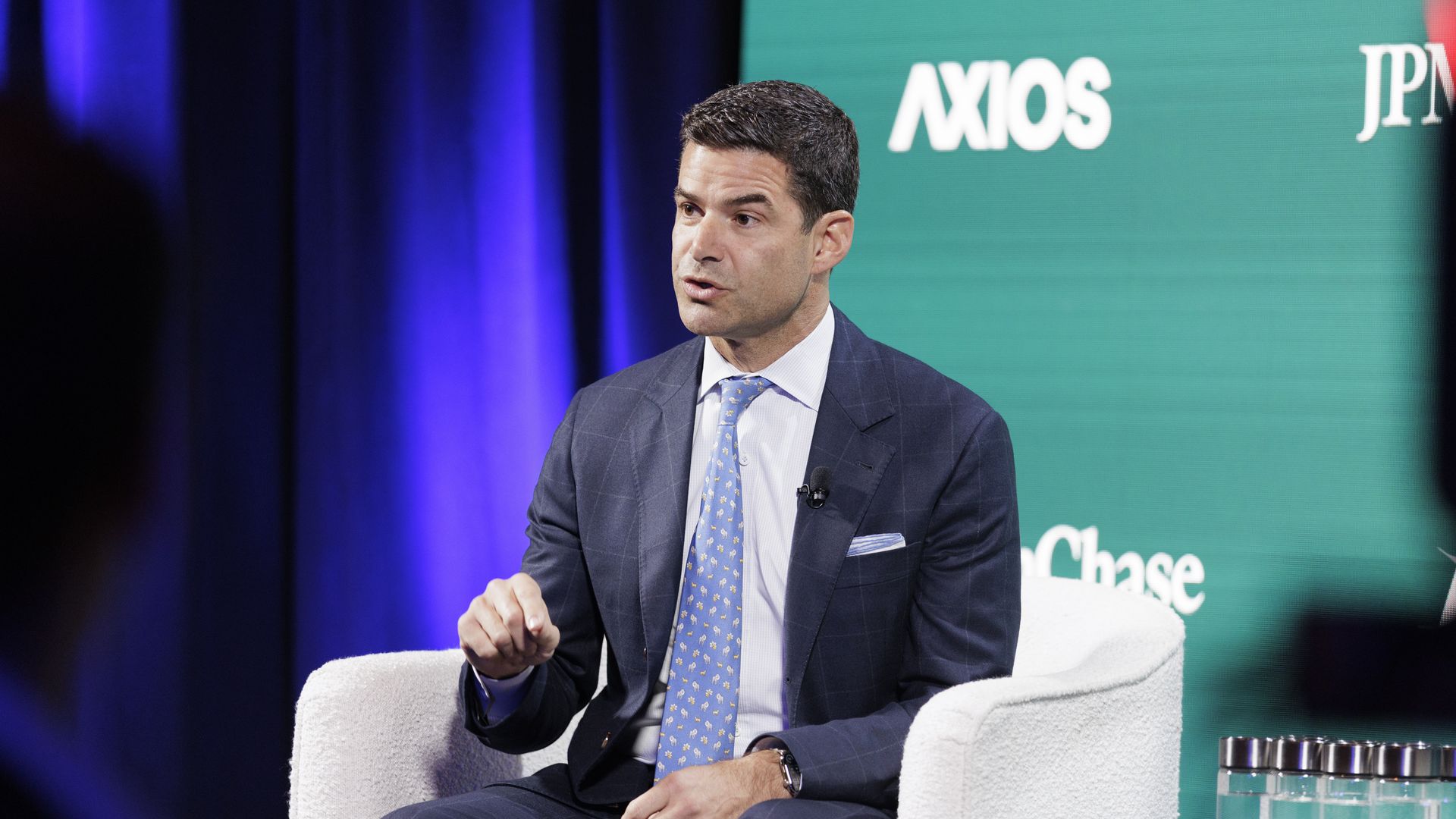 Man in a blue suit and patterned blue tie speaking while seated on a white chair with a microphone on his lapel, purple curtain background, and a green screen with white text behind him.