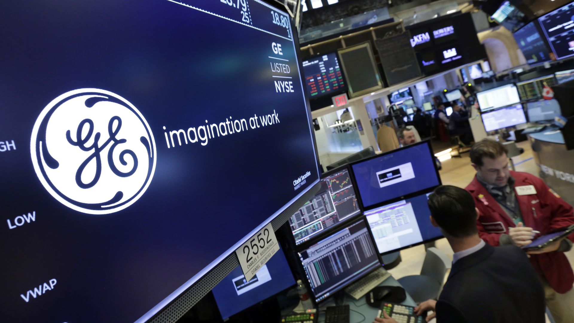GE logo displayed on screens at a stock exchange, with traders in the background
