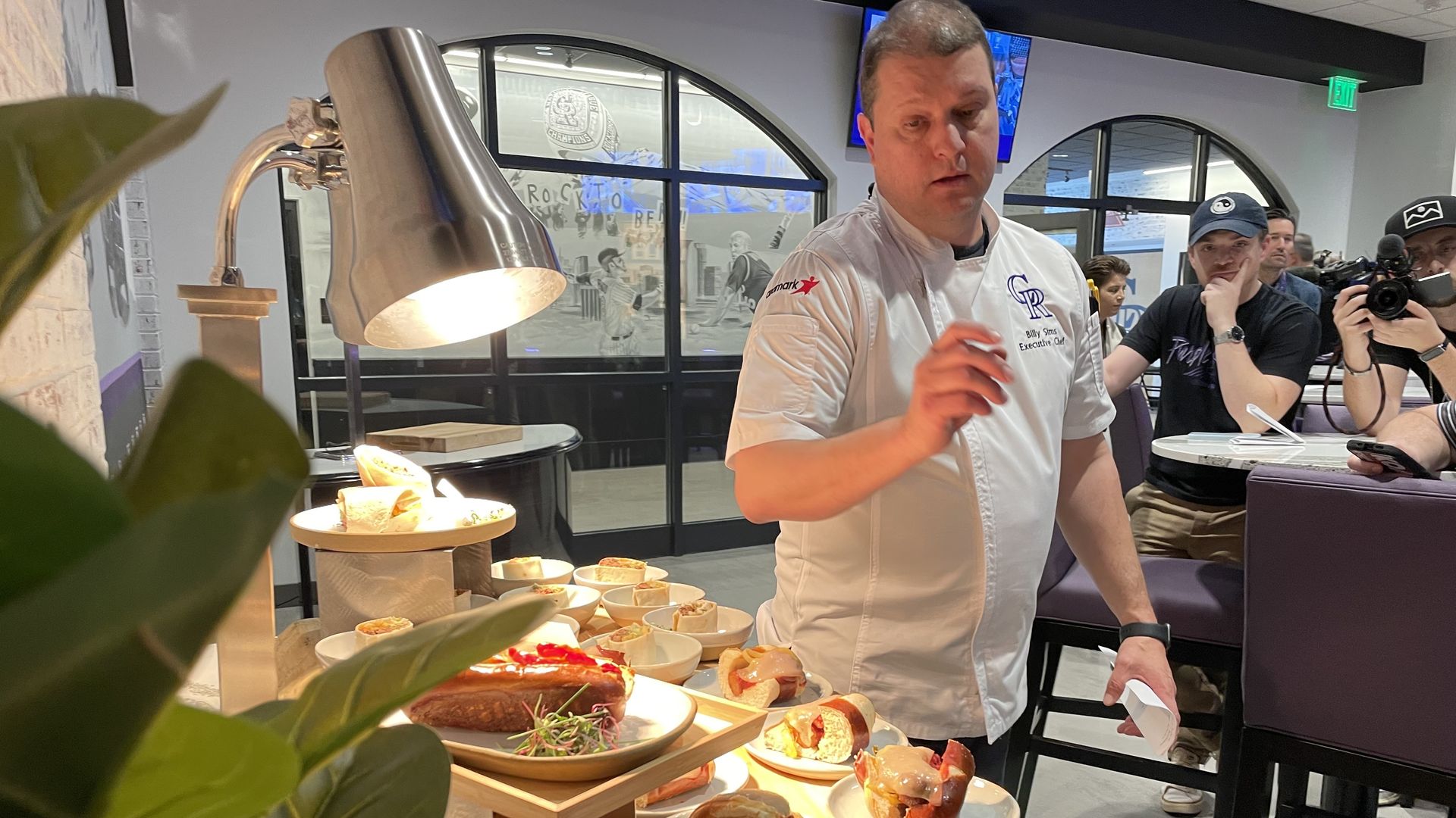A man in a white chefs shirt gestures toward food in several plates in front him, while a light and some plants are in the foreground. Behind them, several people are listening to him speak. 