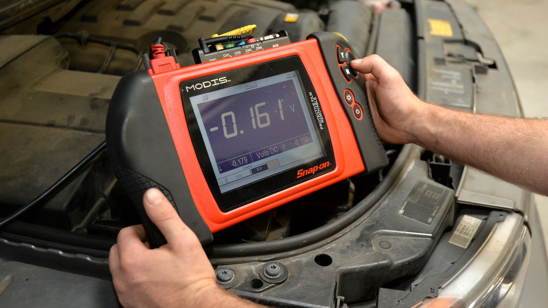 a mechanic holds a diagnostic device with display screen above open car hood and engine block
