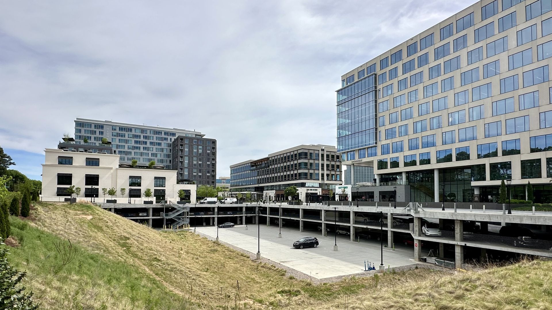 Modern urban scene: a beige glass-front building on the right, mid-rise offices center, a multi-level parking garage, a concrete plaza, and a grassy foreground slope under a blue sky.
