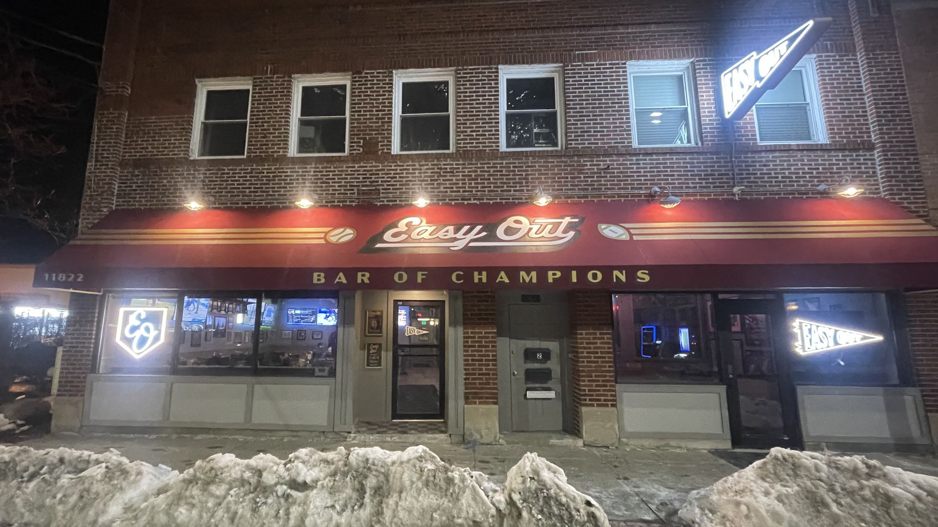 Night view of red-awning bar named Easy Out with signs saying "Bar of Champions" and illuminated logos, snow piled on sidewalk in front of a brick building.