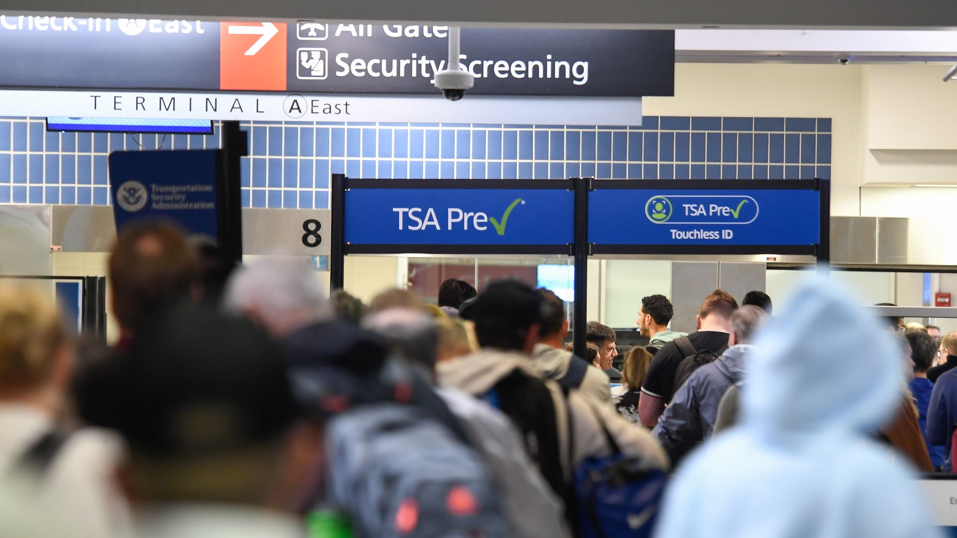 Crowded airport security area with a long line of travelers; blue TSA Pre signs, a Security Screening banner, and Terminal East signs overhead.
