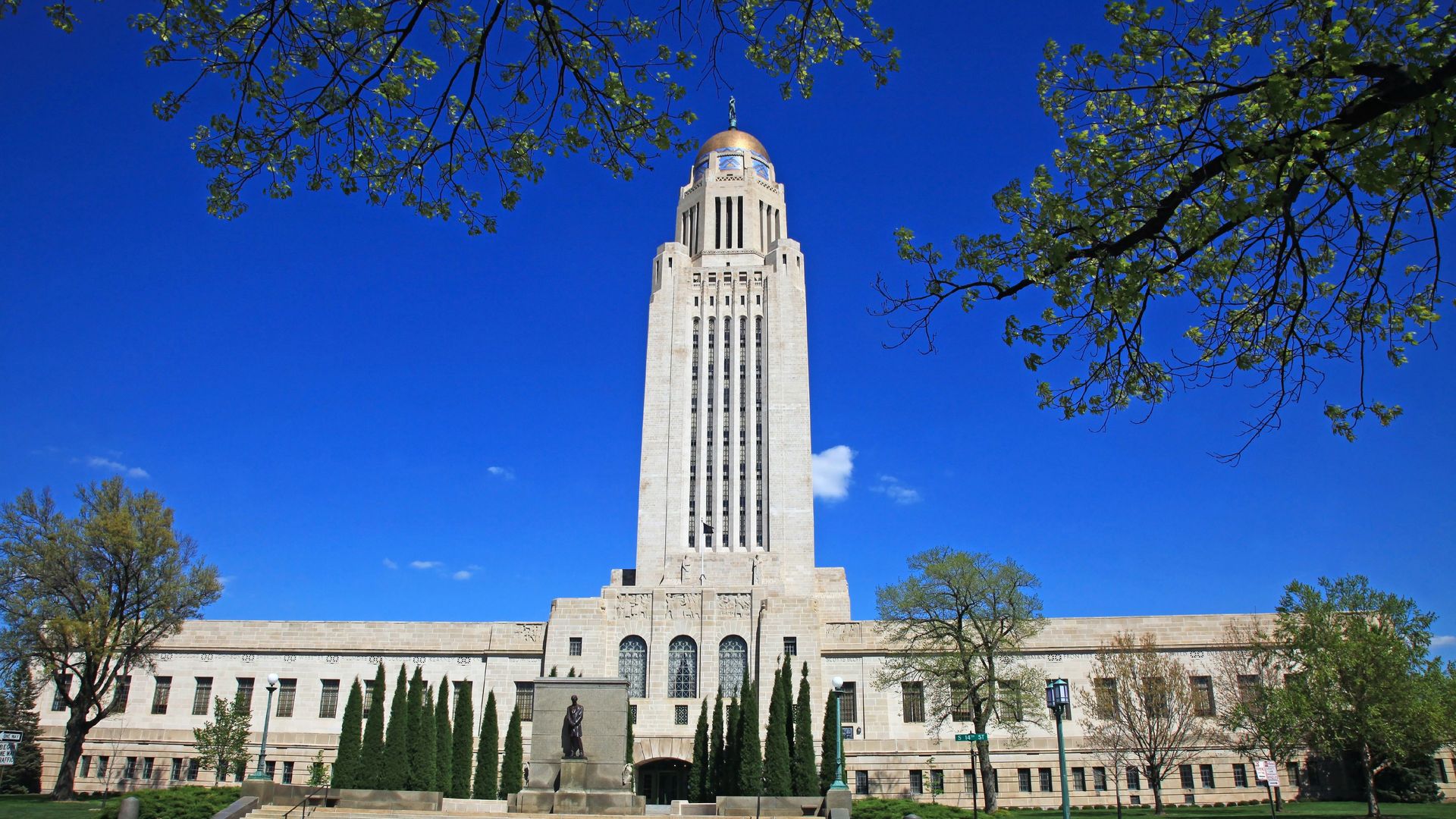An image of Nebraska's state capitol building with a blue sky and trees in the foreground 
