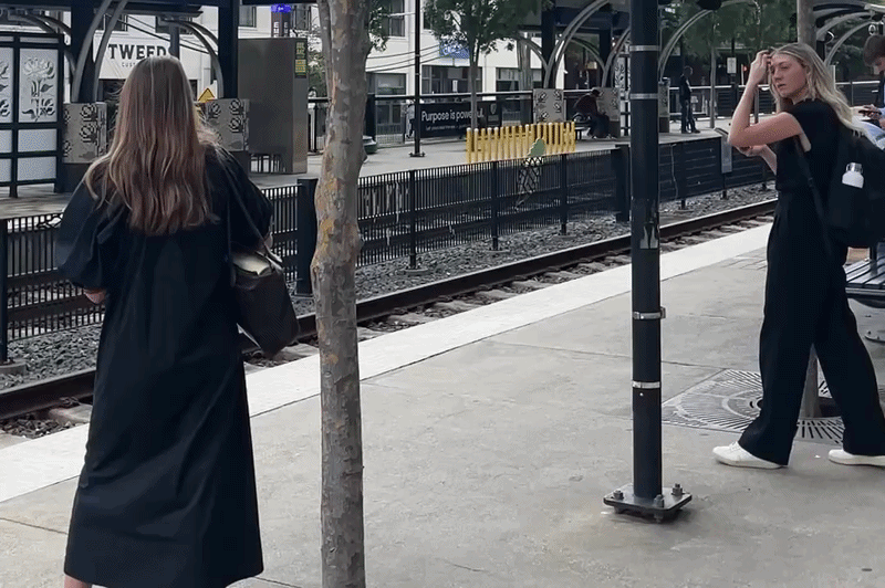 Two women in black outfits on a train platform: one faces the tracks with a shoulder bag, the other adjusts her hair with a backpack; a tree, rails, and yellow safety barricades are in the background.