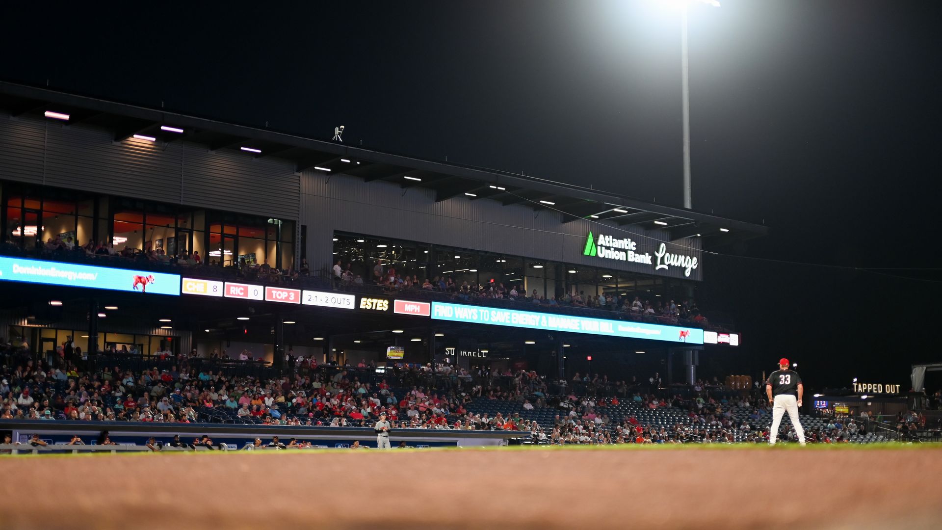Night baseball stadium at night with a tall floodlight illuminating the field. Crowds fill the stands; a sign reads Atlantic Union Bank Lounge while a blue LED scoreboard runs along the front.