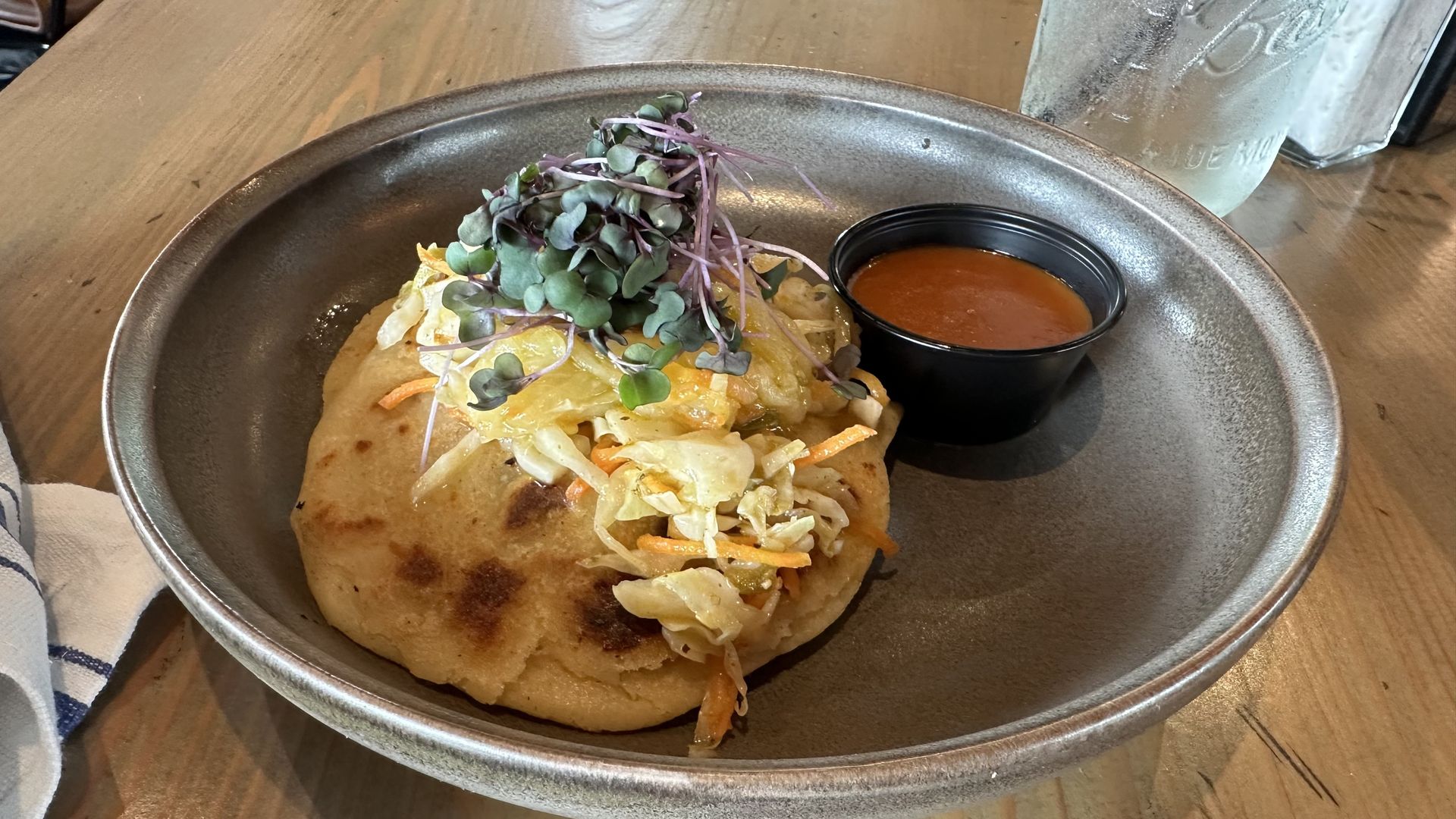 A pupusa sits on a brown plate next to a small container of red sauce.