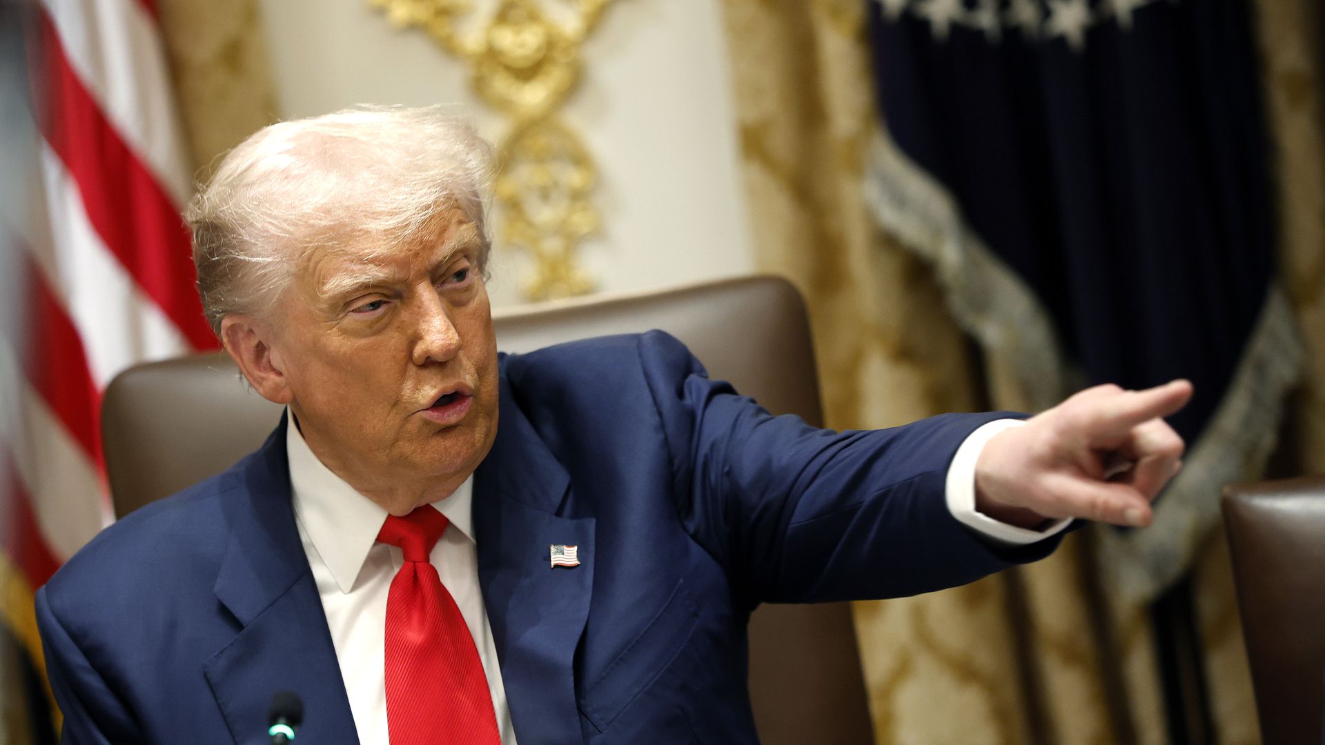 President Donald Trump sits in a leather brown chair and looks to the right, pointing forward with his left hand