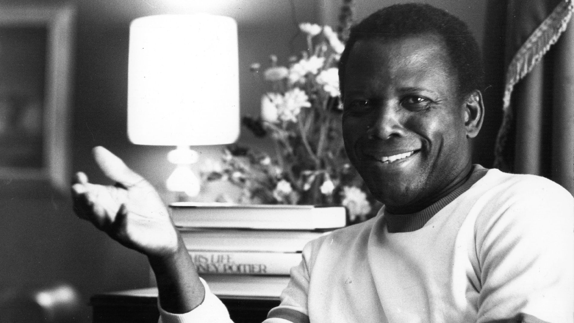 Sidney Poitier, wearing a white sweater, smiles while sitting alongside a bright lamp and extending his right hand.