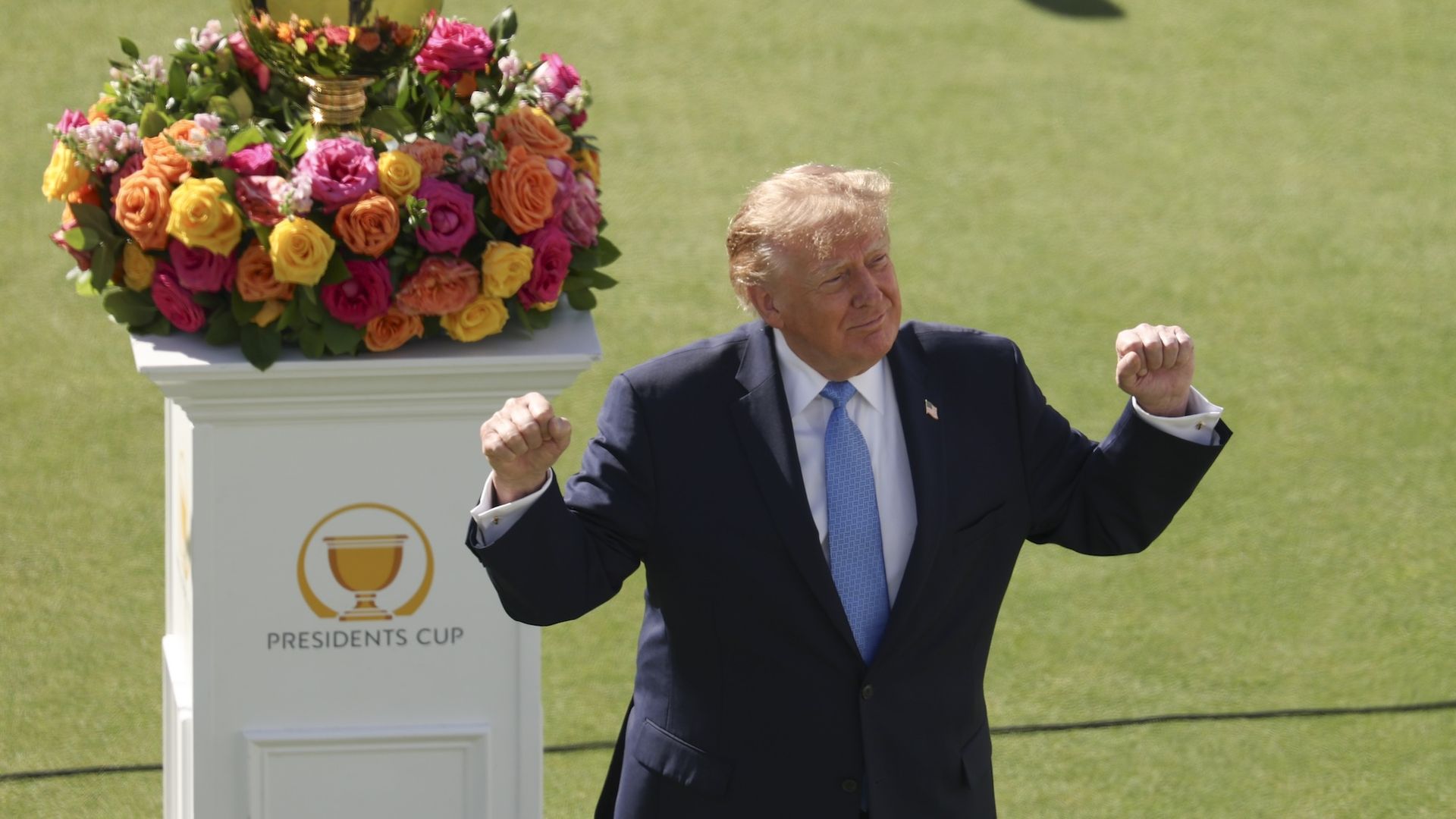 Donald Trump in a dark suit and light blue tie, fists raised, stands beside a podium topped with a gold cup and a vibrant flower arrangement at a Presidents Cup event on a green lawn.