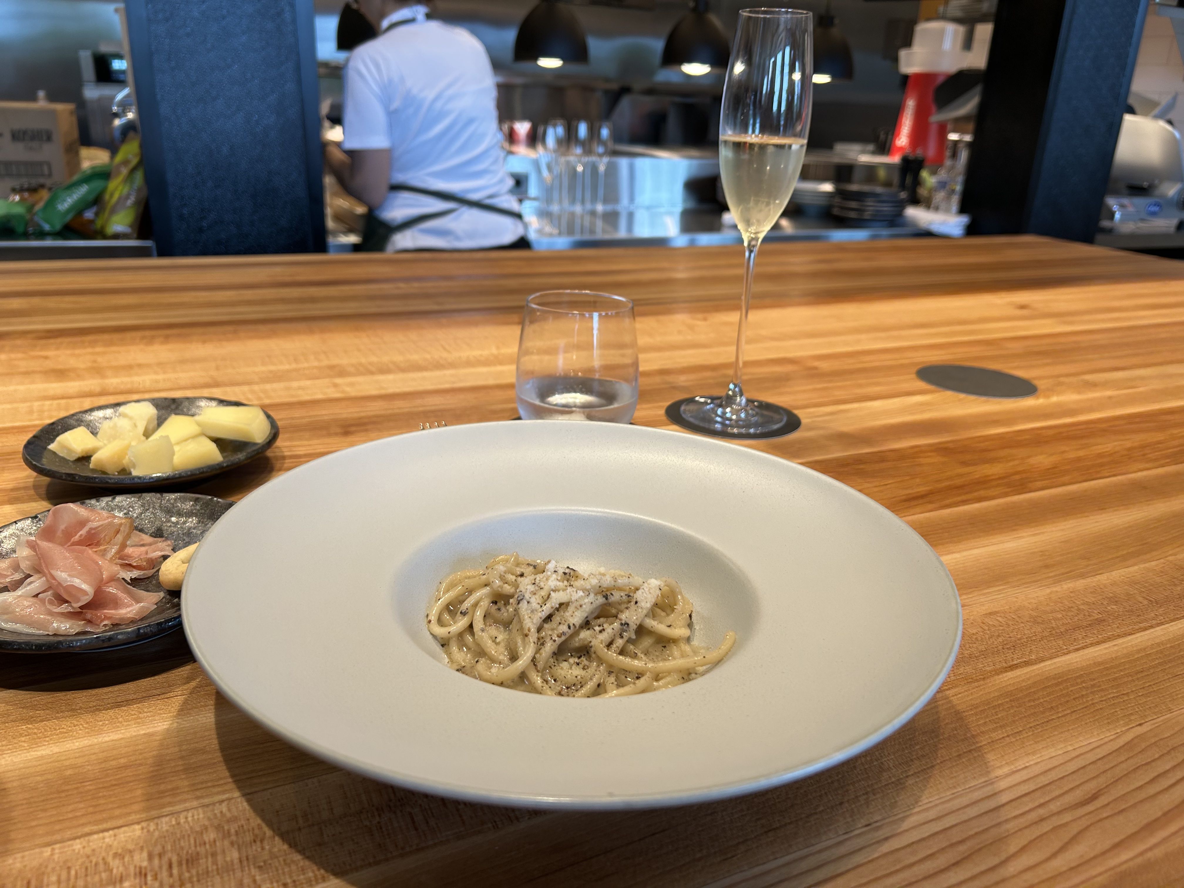 Plate of creamy spaghetti with cheese and pepper on a wooden counter, accompanied by plates of cheese, prosciutto, a glass of water, and a tall glass of sparkling wine.