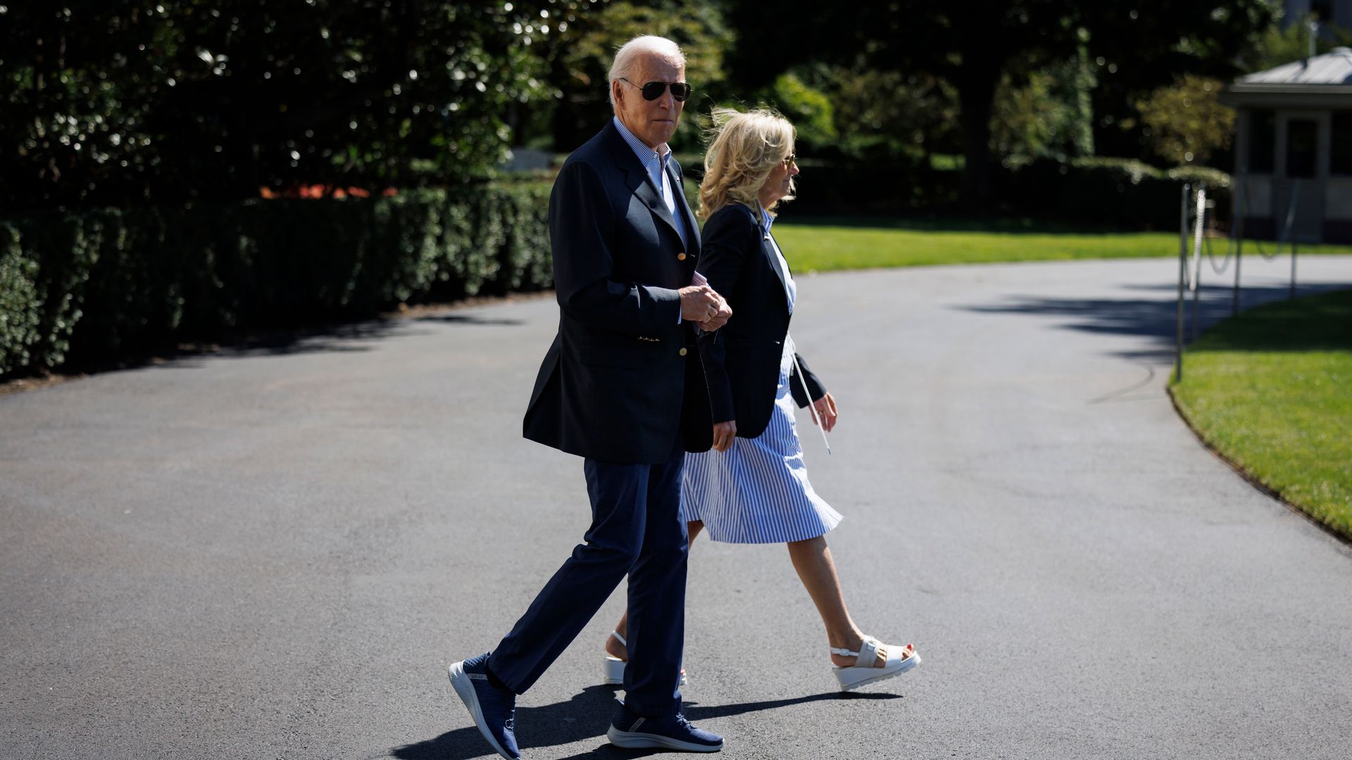 Joe and Jill Biden walk across a driveway at the White House. Joe Biden is wearing a sports jacket, casual trousers and tennis shoes.