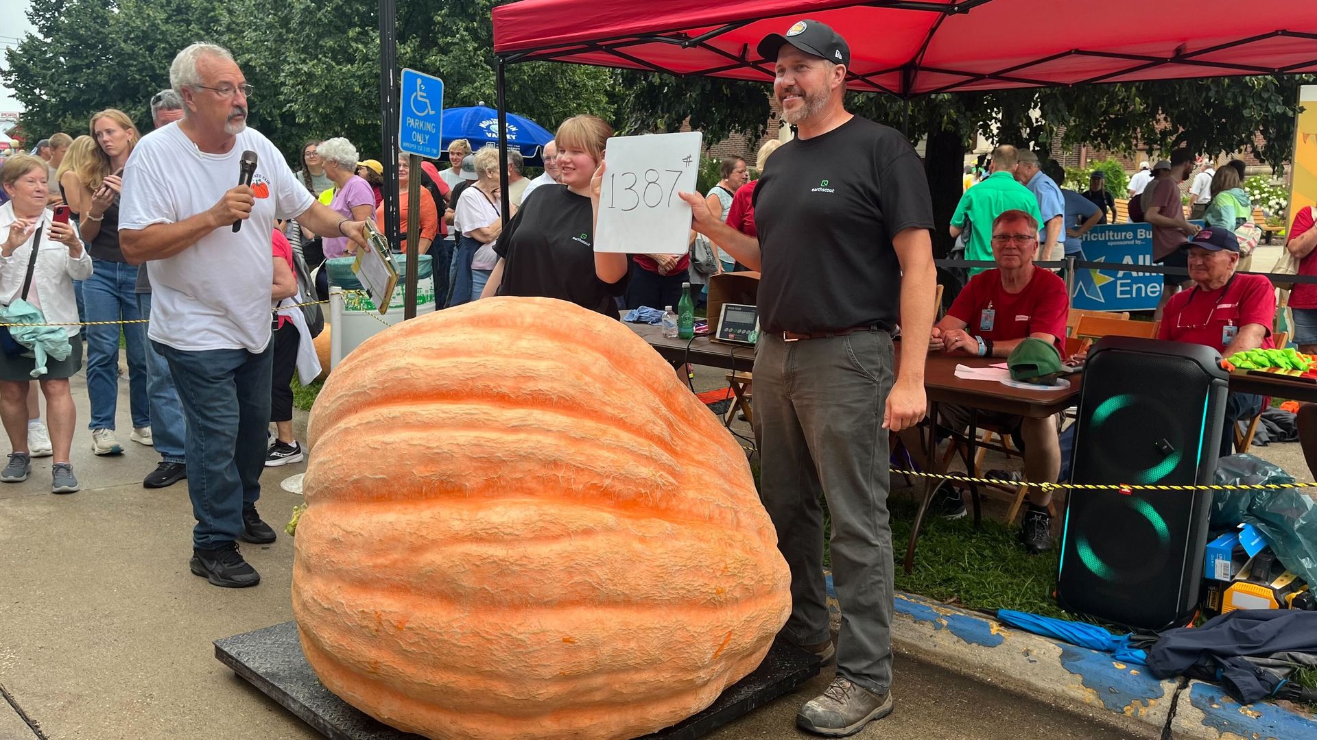 Outdoor festival with a giant orange pumpkin in the foreground. A man in a white shirt speaks into a microphone while another man in black holds a sign reading 1387 under a red tent; crowd and trees in background.