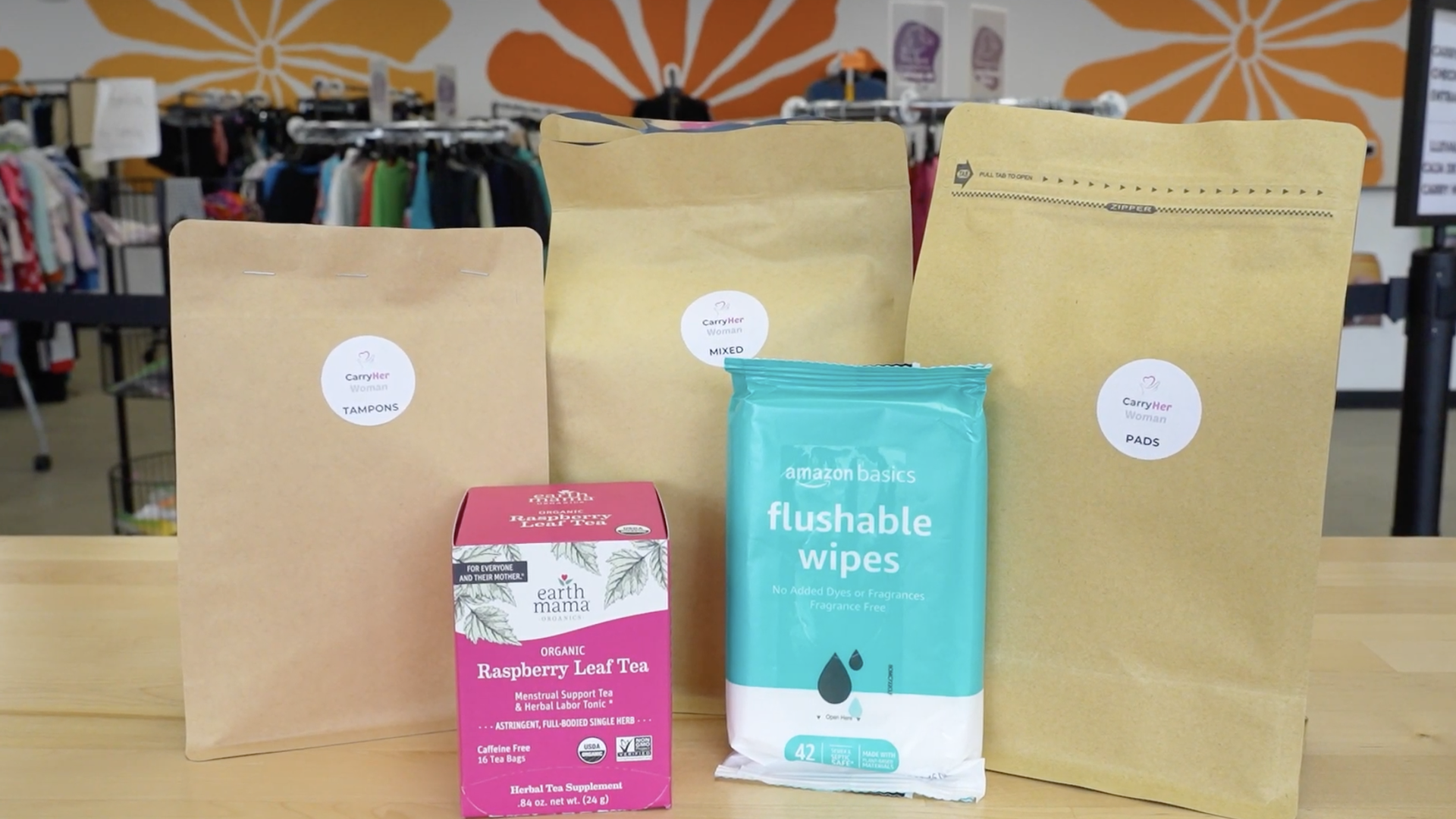 Three large brown bags labeled CarryHer Woman (tampons, pads, mixed) stand on a table with a teal flushable-wipes package and a pink Raspberry Leaf Tea box; blurred clothes racks and an orange floral mural in the background.
