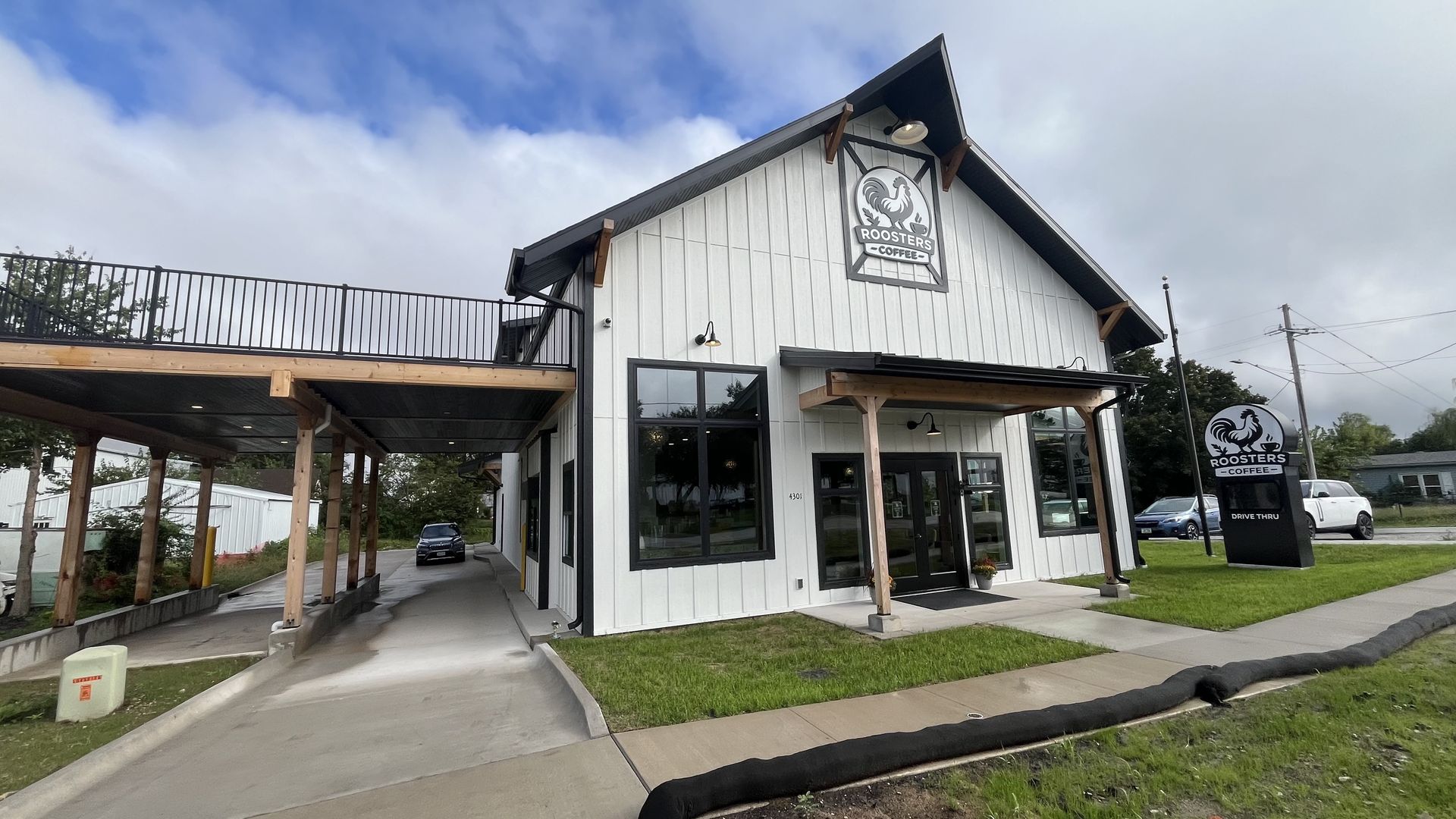 Exterior of a white coffee shop with a gabled roof and Roosters Coffee sign featuring a rooster logo. A wooden-covered walkway lines the side, large front windows, and a drive-thru sign nearby.