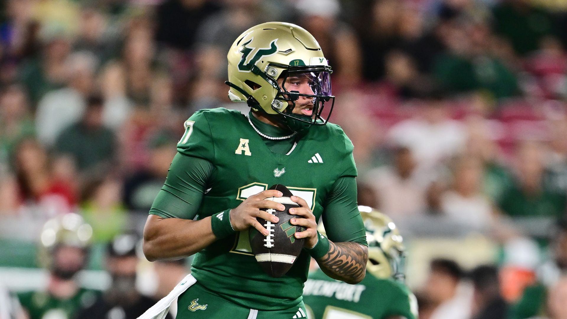 Football player in green and gold USF Bulls uniform holding ball ready to throw, with helmet and dark arm tattoo, blurred crowd in background.