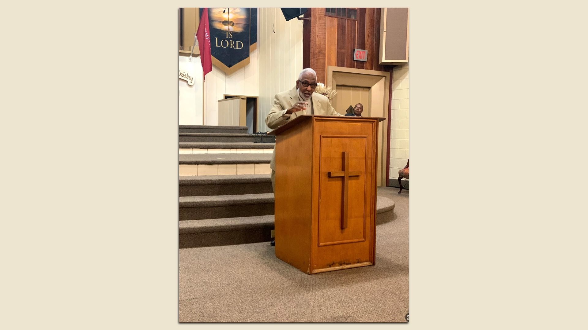 Man in beige suit speaking at wooden pulpit with cross in church setting, banner reading "IS LORD" in background, and man sitting by door.