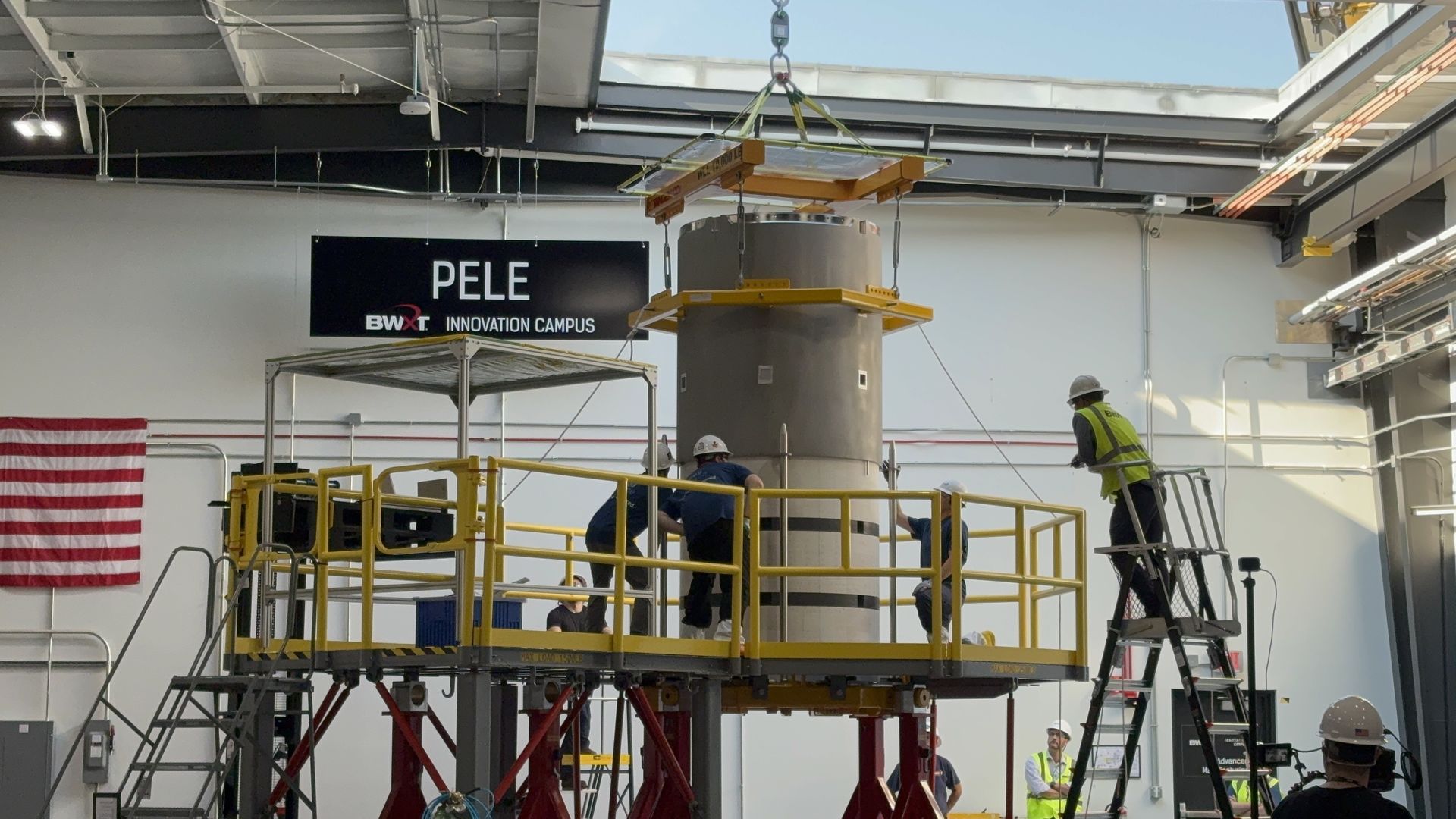 Inside a bright industrial hangar, a large cylindrical vessel is suspended by a crane with yellow scaffolding. Workers in hard hats observe from a platform near a sign.
