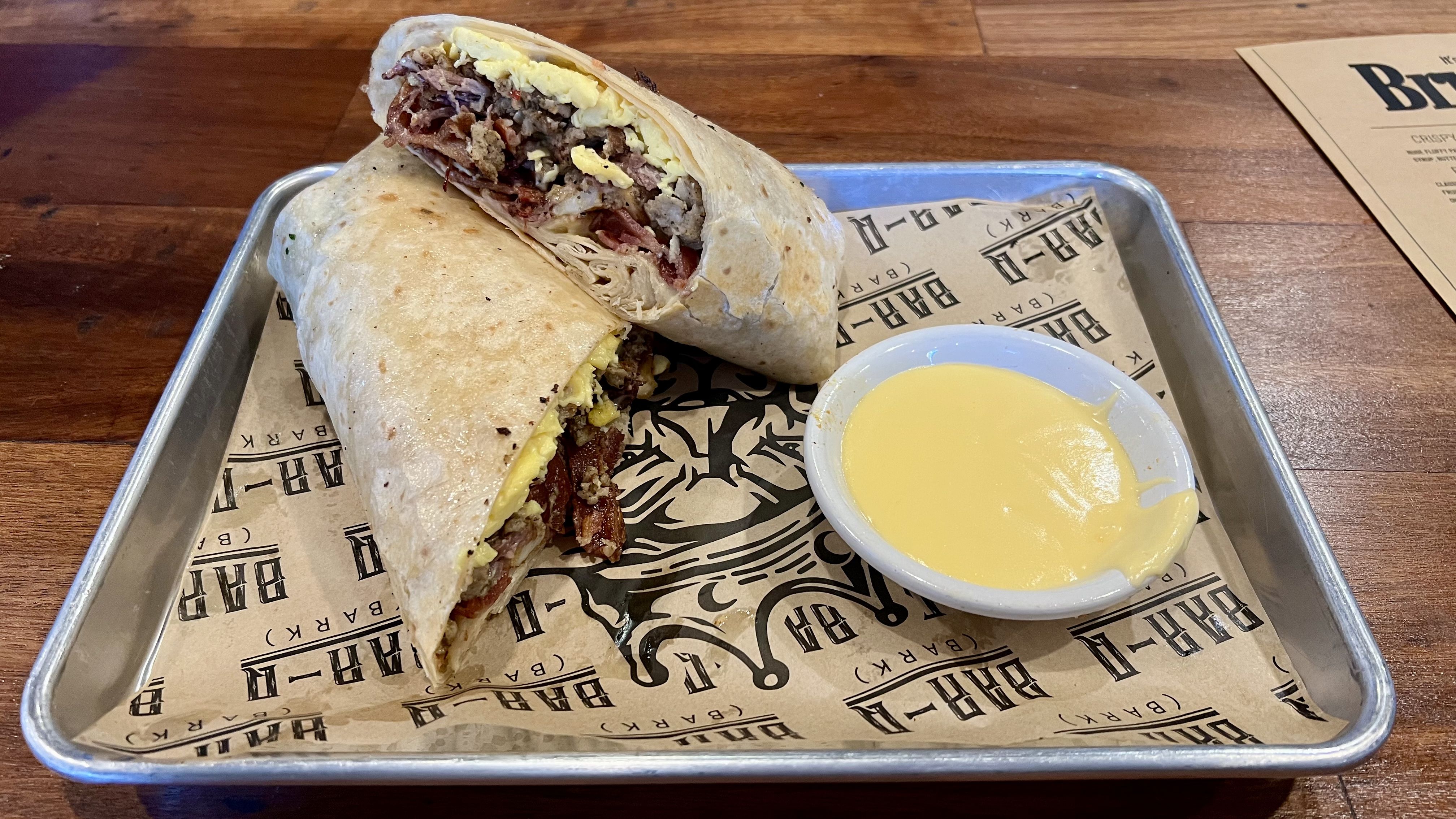 Breakfast burrito cut in half with eggs, sausage, and bacon on branded paper in a metal tray with a side of yellow cheese sauce on a wooden table.