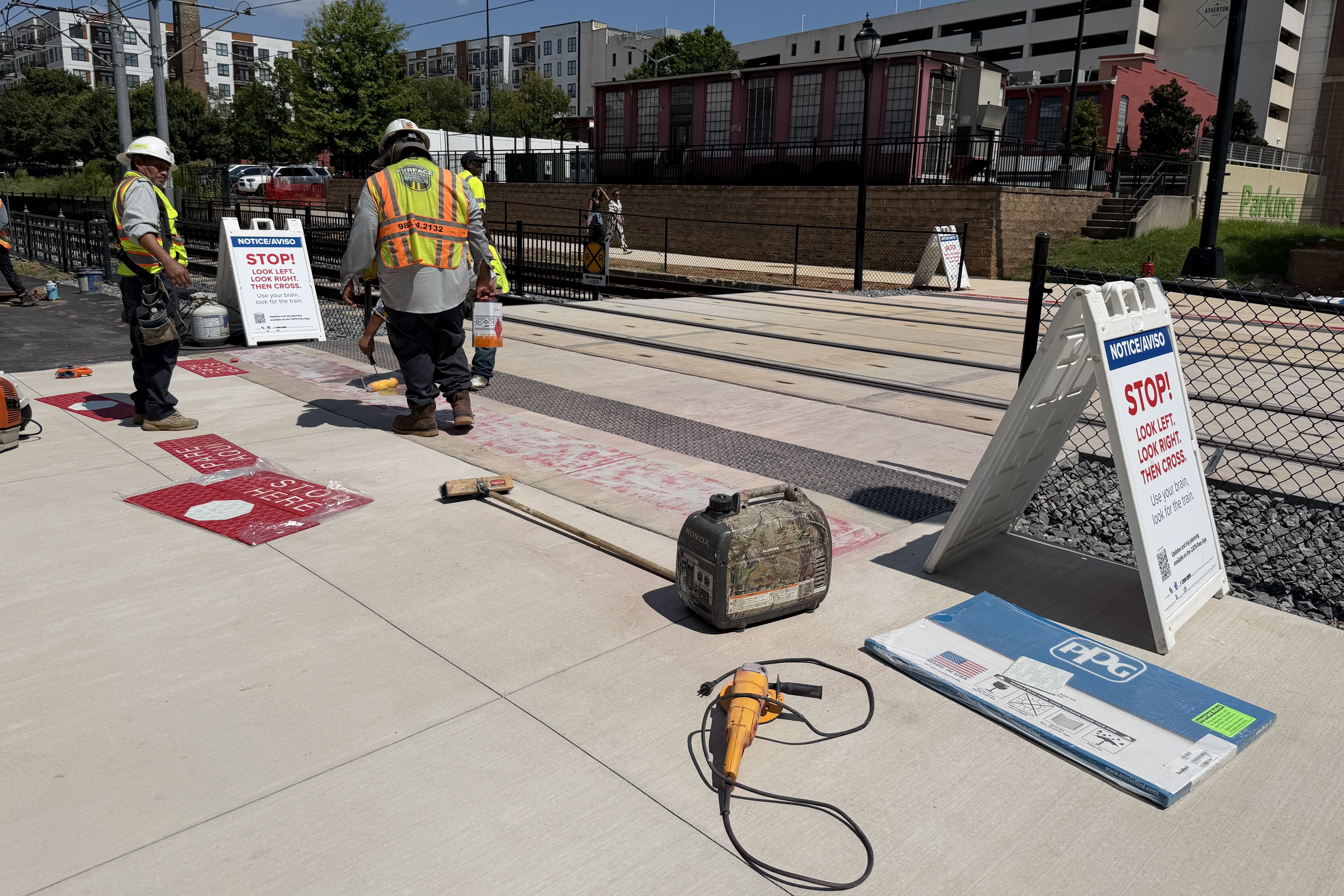 Construction workers in hi-vis vests and hard hats stand beside a fenced railroad crossing. Signs read STOP and LOOK LEFT, LOOK RIGHT, THEN CROSS; urban buildings and equipment visible in the background.