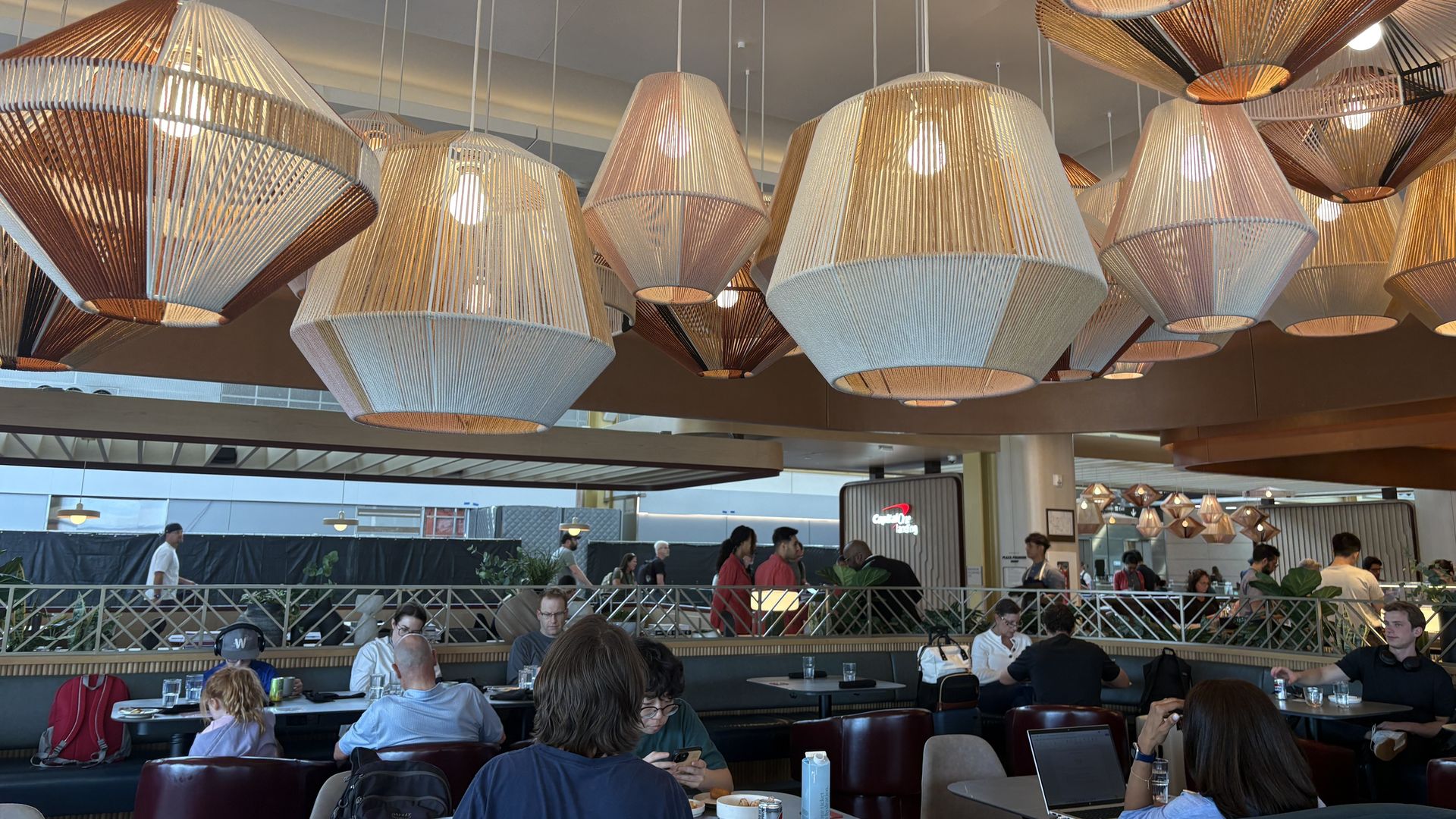 Interior of a busy cafe with dozens of woven pendant lamps in cream, tan, and brown hues hanging from the ceiling; customers sit at tables, some with laptops and drinks.
