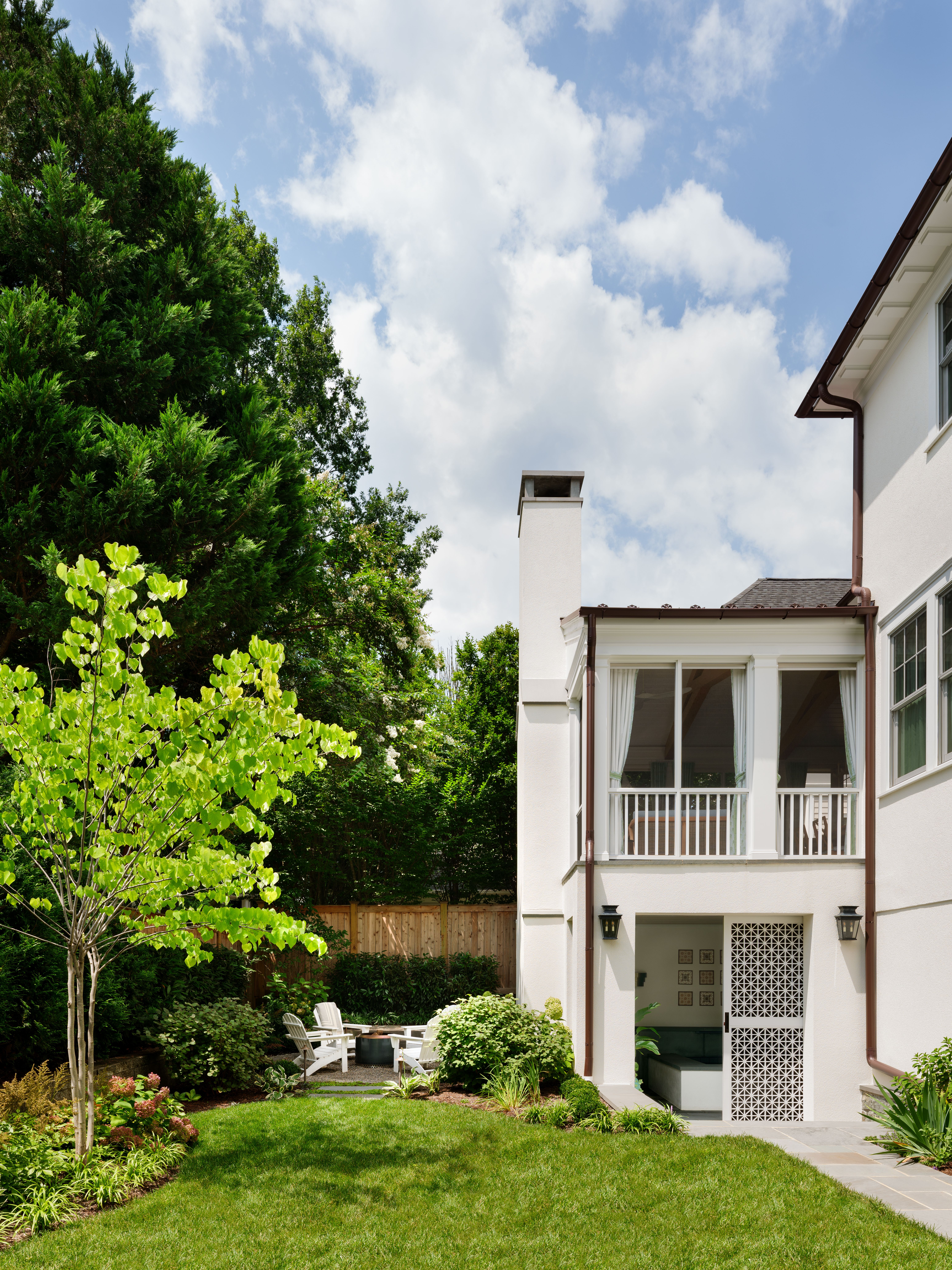 A sunny backyard shows a two-story white house with a porch on the right, a chimney and a glass door. Green lawn, tall trees, a wooden fence, and a seating area with white chairs around a fire pit.