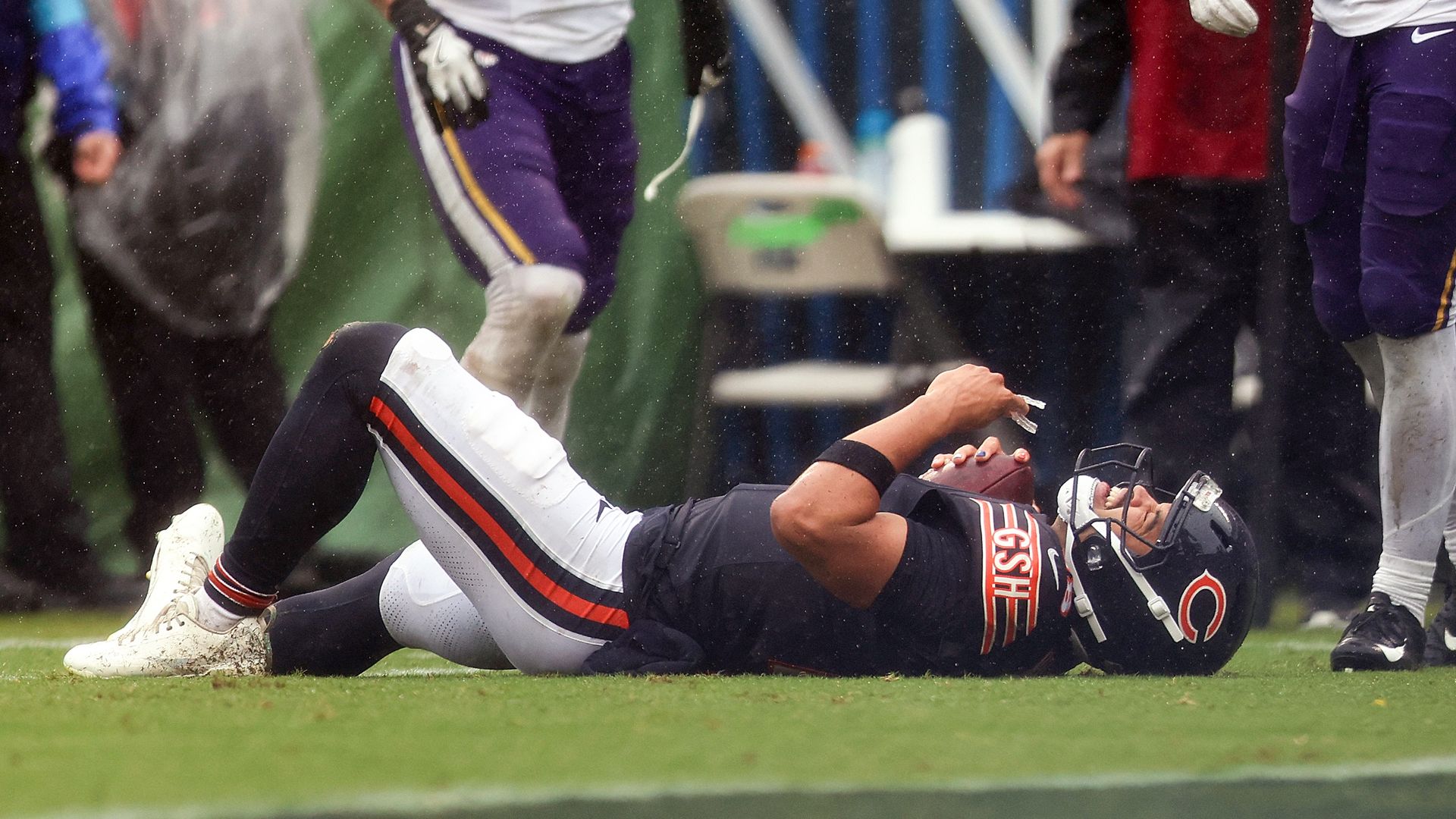 Football scene on a rainy day: Caleb Williams in a black uniform with red trim lies on the grass with helmet on, while teammates in white jerseys and purple pants stand nearby amid raindrops.