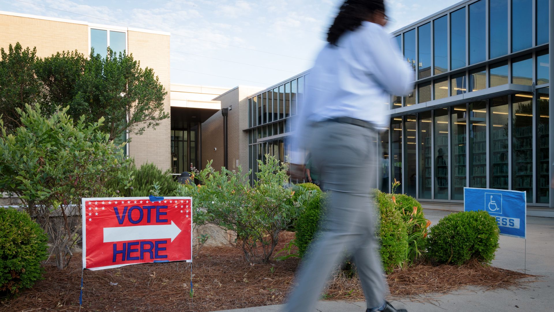 A red sign that reads" vote here" in blue lettering, with an arrow pointing to the right, is perched into the ground outside a modern building with many windows. A person wearing gray pants, a light blue long-sleeved shirt and black shoes walks by in the foreground. 