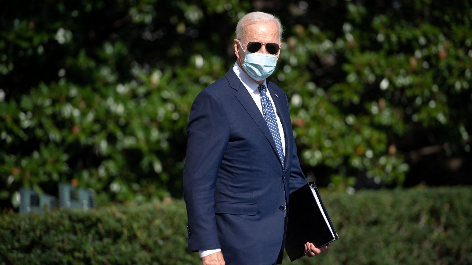 Photo of Joe Biden wearing sunglasses and a mask as he walks with a binder in one hand