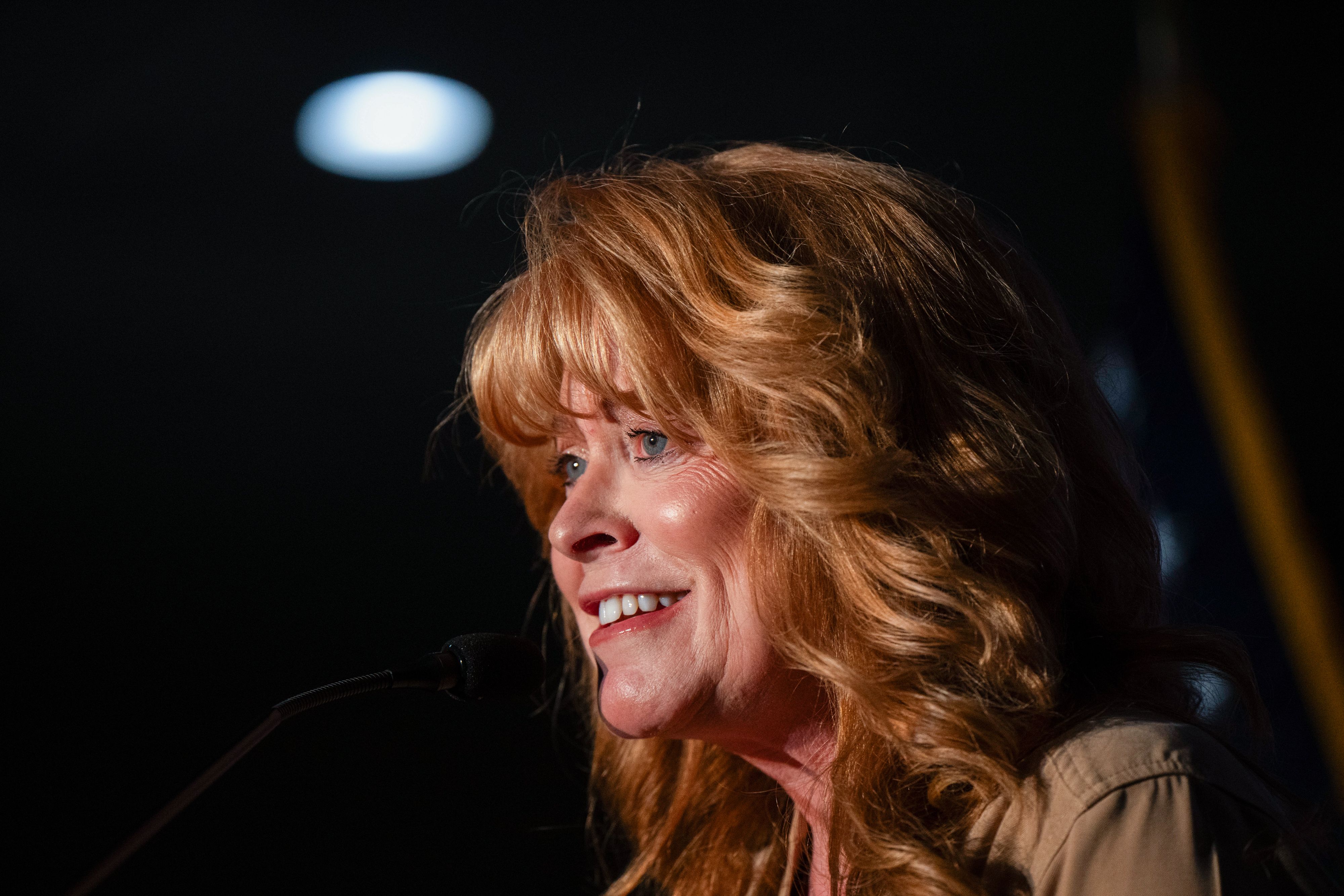 Close-up of a smiling woman with light brown hair speaking into a microphone on a dark stage. Warm lighting highlights her face and hair, with a blurred blue light in the background.