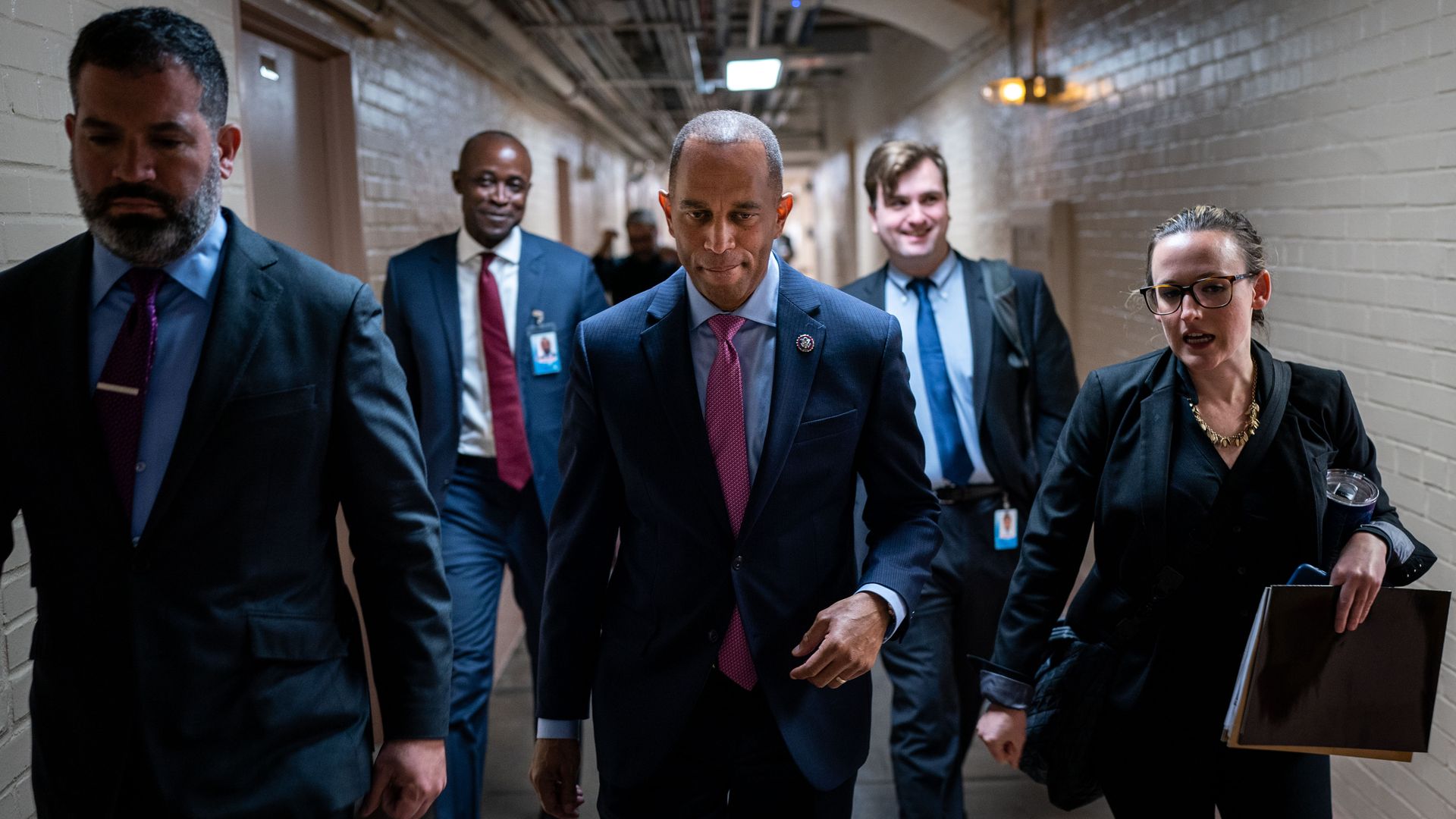 Rep. Hakeem Jeffries, wearing a blue suit jacket, light blue shirt and red tie, walks in a Capitol basement hallway flanked by staff and security.
