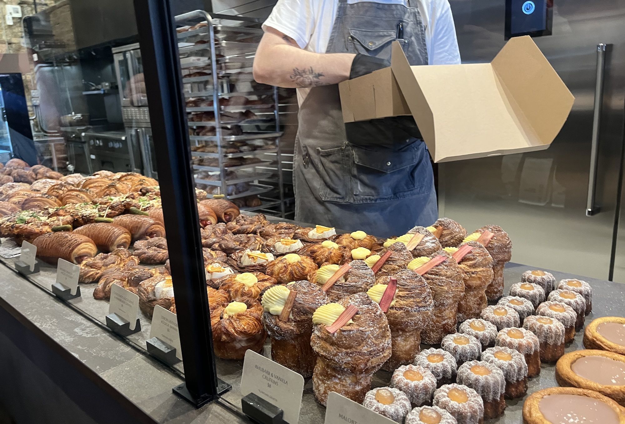 Bakery display window full of croissants and pastries; worker in a light shirt and gray apron holds an open cardboard box, wearing a glove, with racks of trays behind.