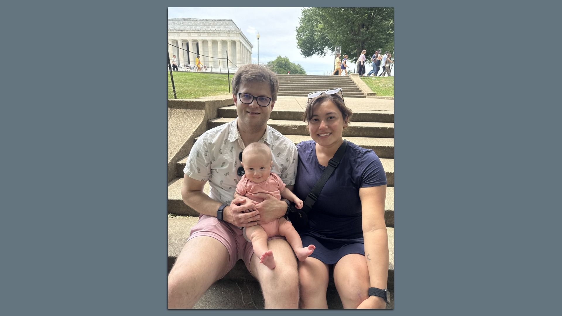 A family of three sits on outdoor stone steps in front of a grand neoclassical building. The man with glasses holds a baby in pink while the woman smiles beside him.