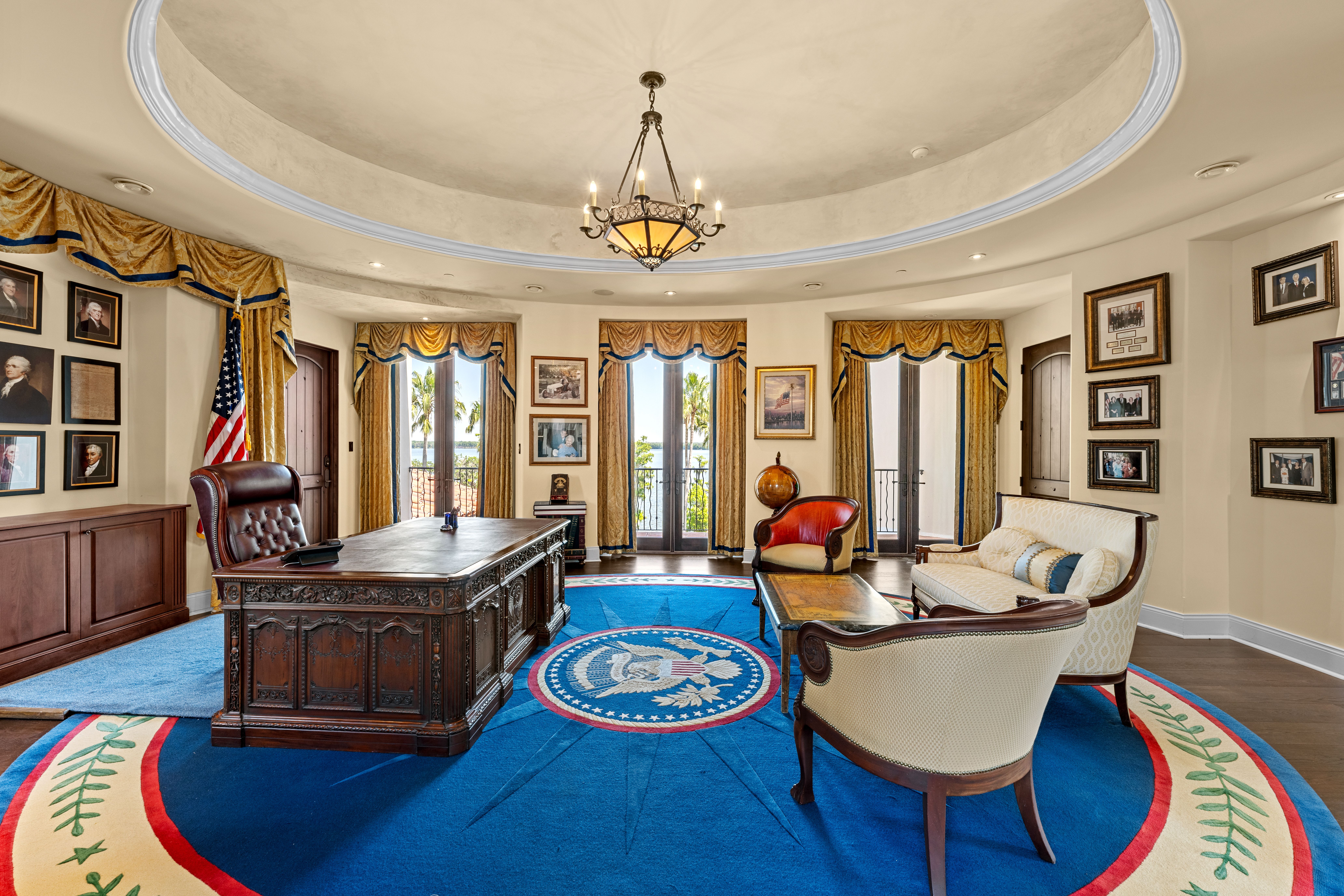 Elegant oval office-inspired room with large carved wooden desk, blue carpet with presidential seal, beige and red chairs, gold curtains, American flag, and photos on cream walls.