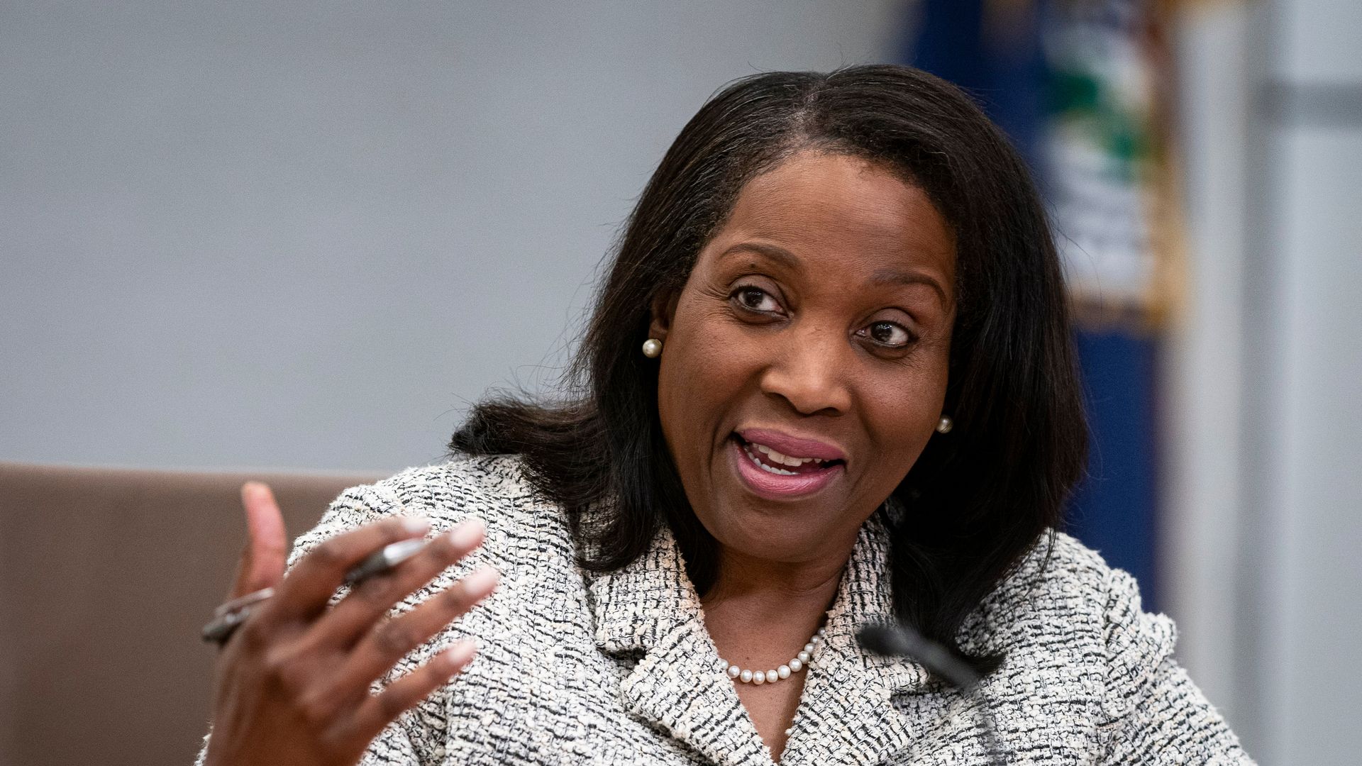 Woman with medium-dark skin, black shoulder-length hair, wearing pearl earrings and necklace and a white textured jacket, speaking and gesturing with a pen in her hand.