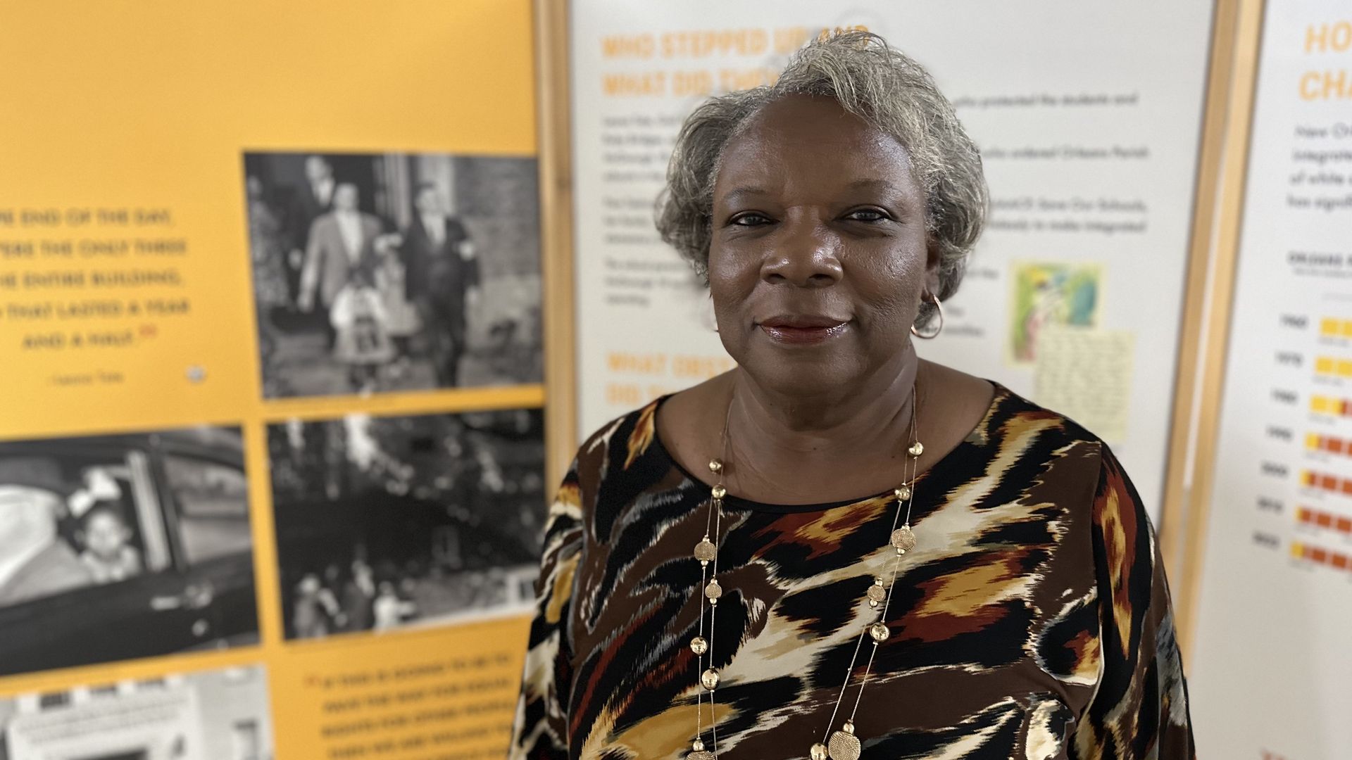 Leona Tate stands in front a museum panel telling the story of how she was part of the desegregation of New Orleans schools. She looks directly in the camera and smiles slightly.