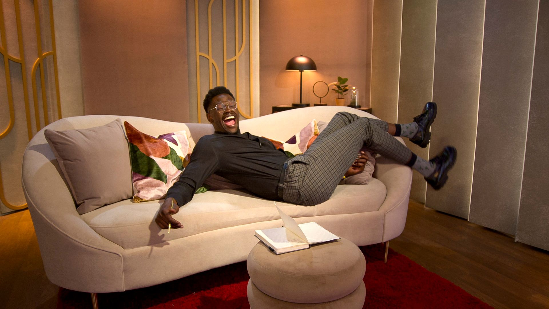 A joyful man wearing glasses, black shirt, and checkered pants is laughing and lying back on a beige couch with colorful cushions in a warmly lit room with modern decor.