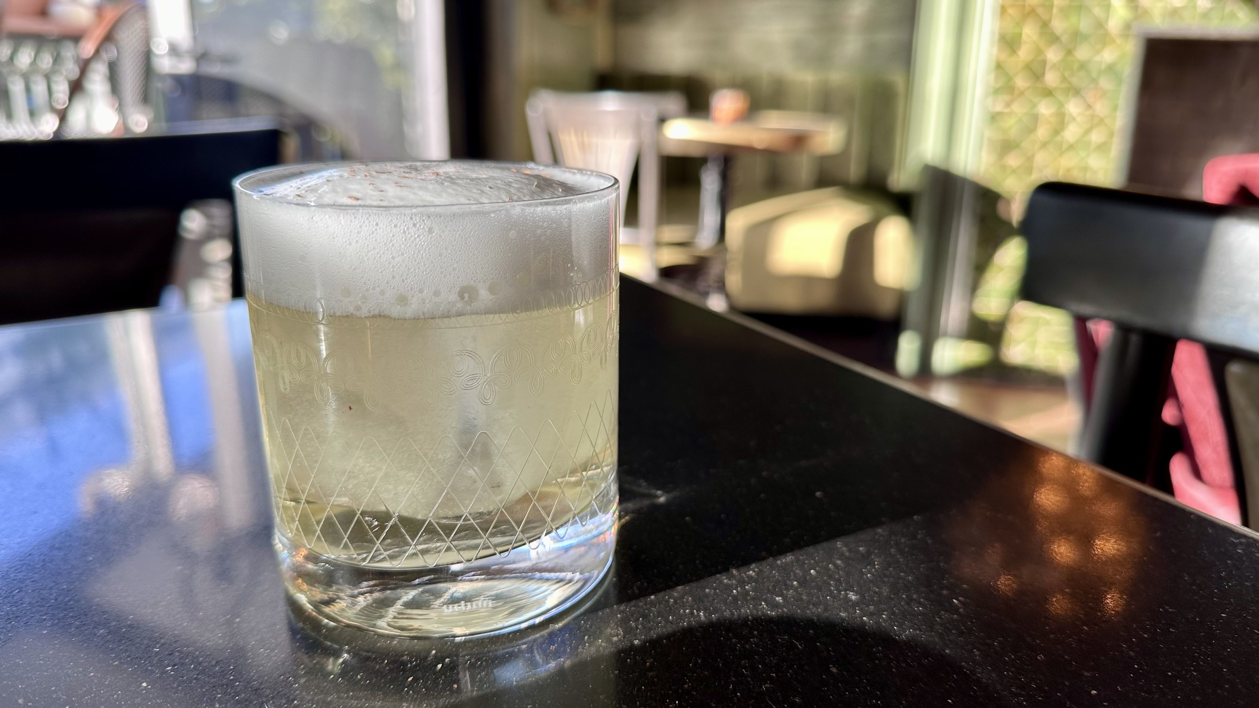 Clear glass with etched patterns containing a pale yellow frothy drink and ice cubes, sitting on a shiny black table in a softly lit cafe with chairs and tables blurred in background.