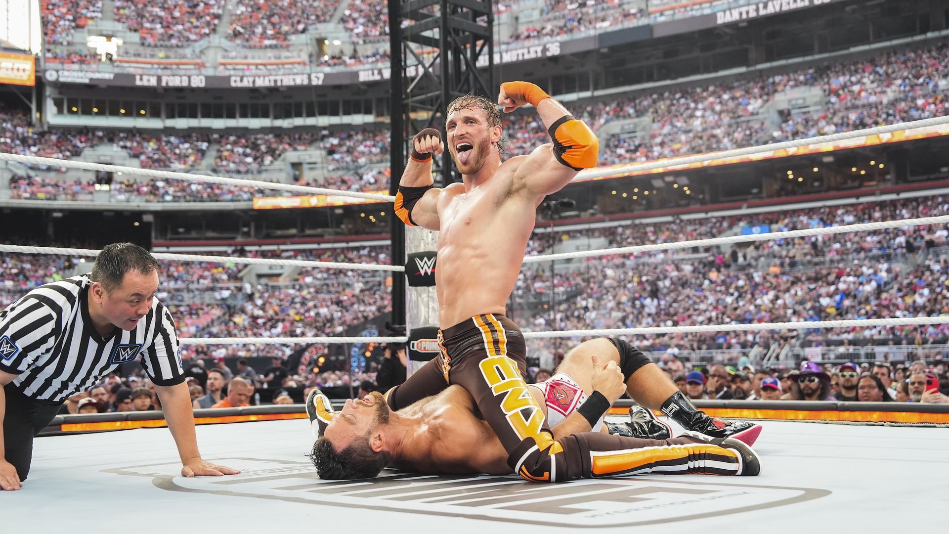 Logan Paul does a split while flexing on WWE star L.A. Knight in a ring at a stadium. 